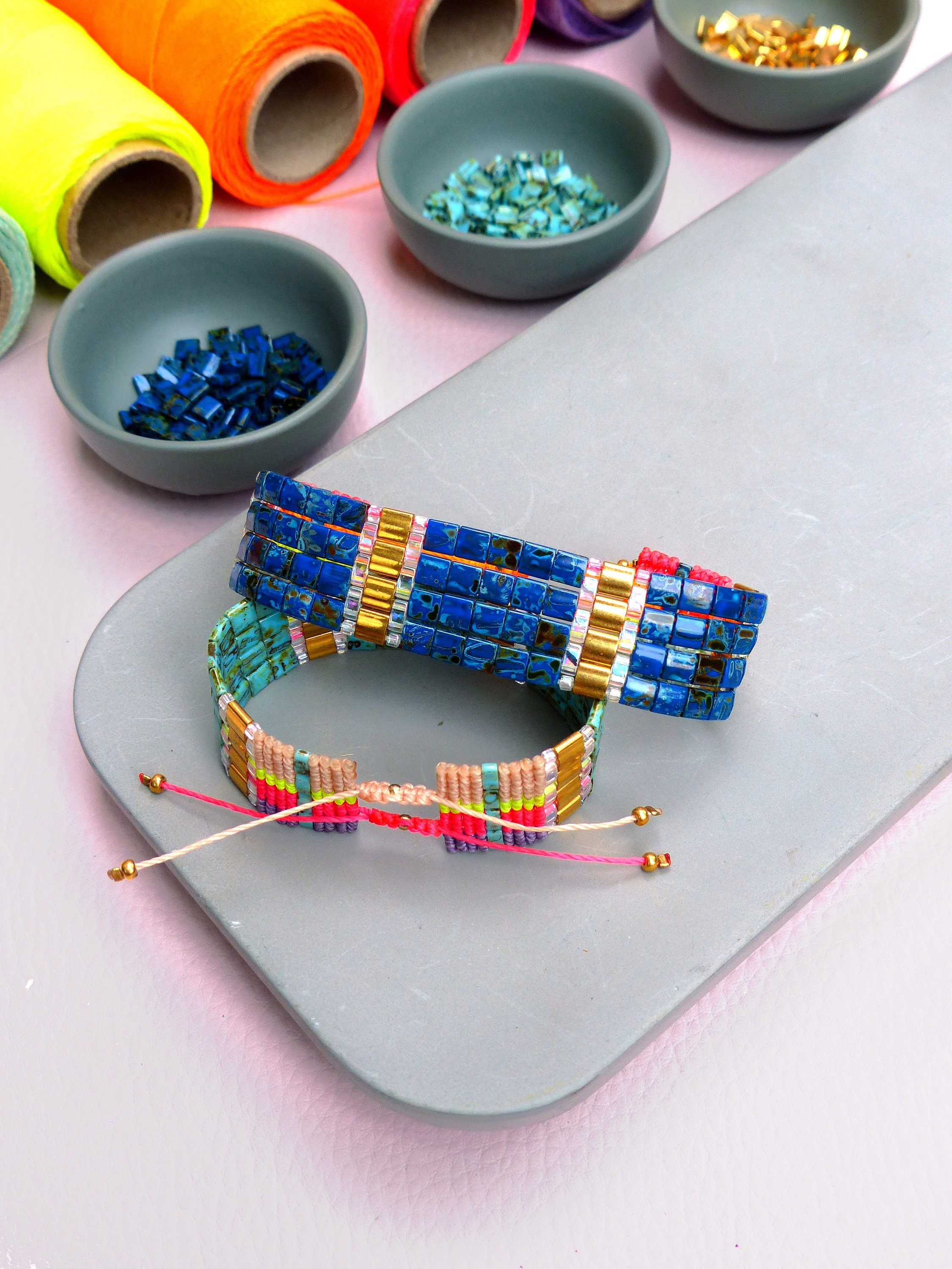 Peut inclure: Deux bracelets en perles colorées sur une surface grise. Le bracelet du haut est bleu avec des accents dorés et le bracelet du bas est multicolore avec des fils roses, jaunes et verts.