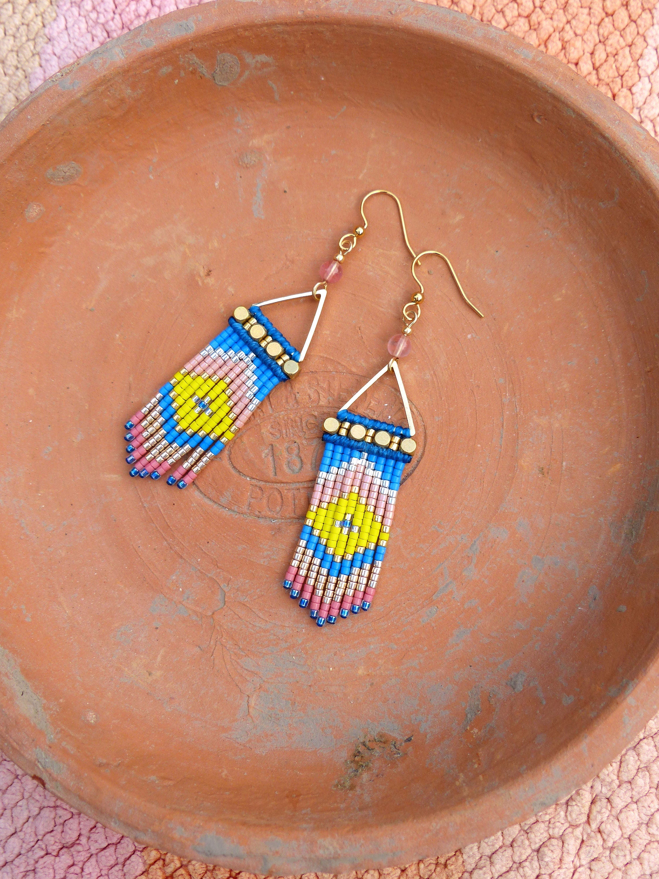 Peut inclure: Boucles d'oreilles en perles colorées avec un motif géométrique. Elles présentent un haut triangulaire doré et une frange de perles bleues, roses, jaunes et argentées. Posées sur une assiette en terre cuite.
