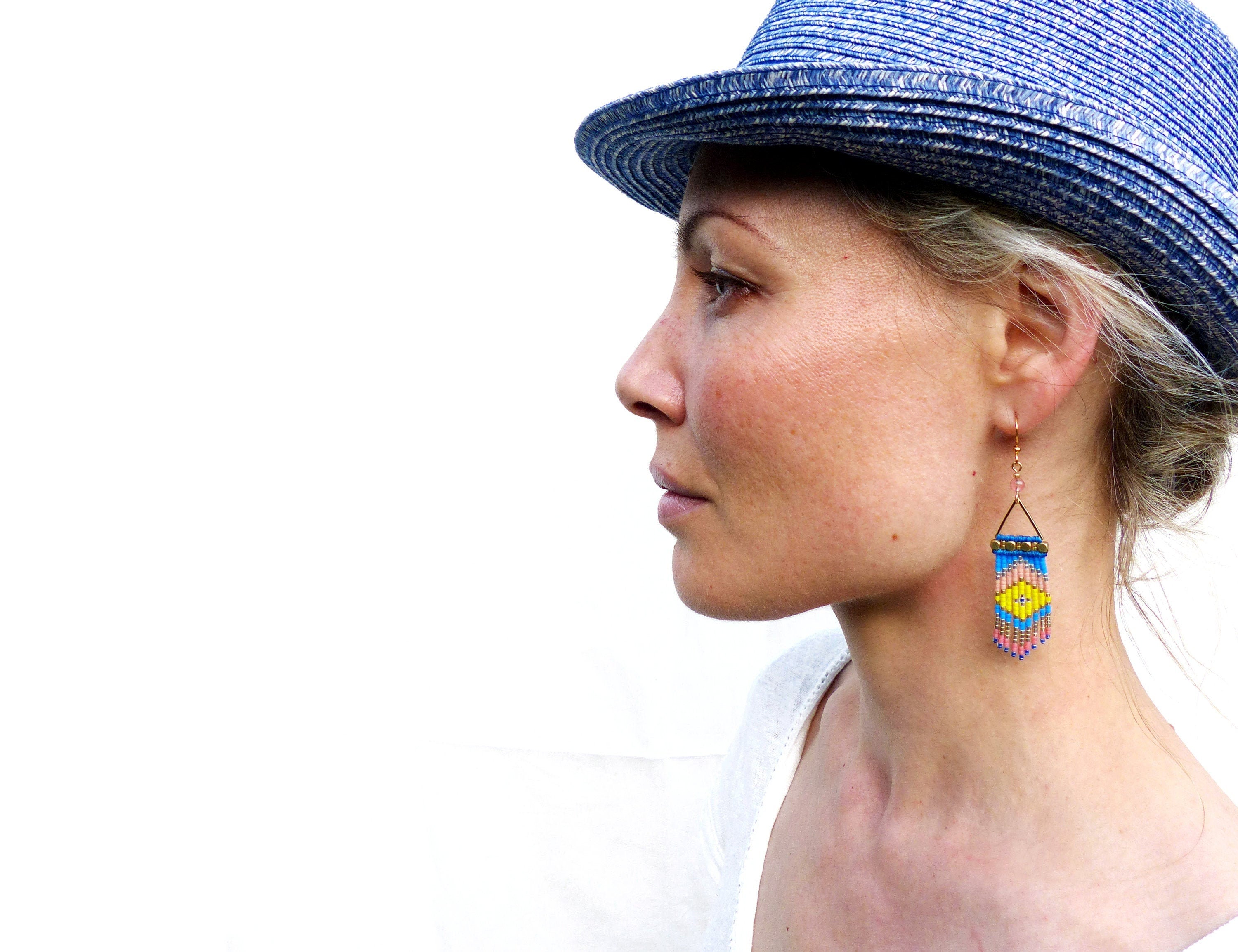 Peut inclure: Profil d'une personne portant un chapeau fedora bleu et des boucles d'oreilles dorées. Les boucles d'oreilles sont rectangulaires avec un motif géométrique en perles bleues, jaunes, roses et noires. Les boucles d'oreilles sont suspendues à des crochets dorés.
