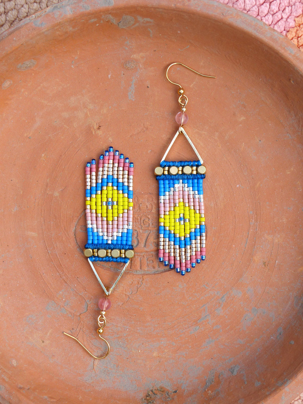 Peut inclure: Une paire de boucles d'oreilles perlées faites à la main avec un motif géométrique. Les boucles d'oreilles présentent une combinaison de perles bleues, roses, jaunes et dorées, disposées en forme rectangulaire avec un cadre triangulaire doré et des crochets d'oreilles dorés.