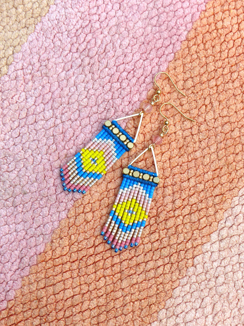 Peut inclure: Paire de boucles d'oreilles en perles avec un motif géométrique bleu, jaune et rose. Les boucles d'oreilles ont des crochets et des accents dorés. Elles sont présentées sur une surface texturée multicolore.