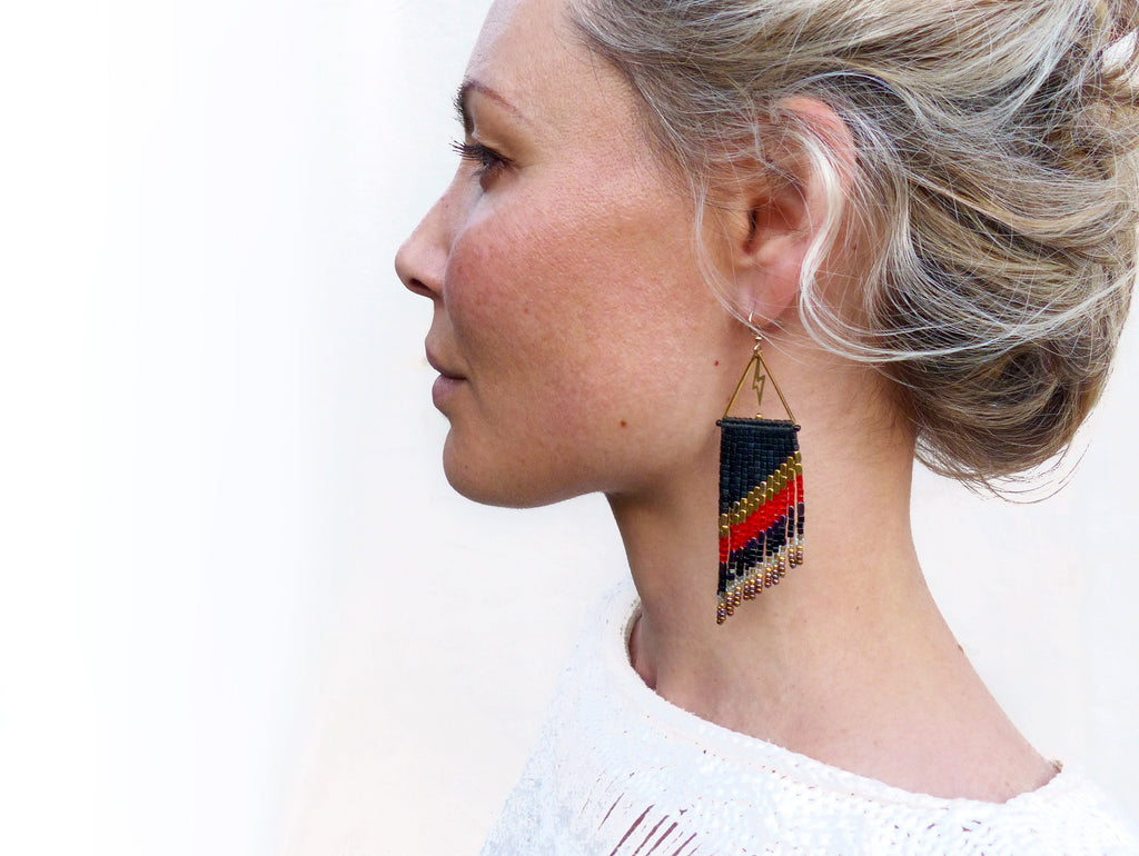Peut inclure: Boucles d'oreilles perlées faites à la main avec un motif géométrique. Les boucles d'oreilles présentent une base noire avec des rayures diagonales rouges et dorées, et des franges dorées. Elles sont suspendues à des cadres triangulaires dorés.