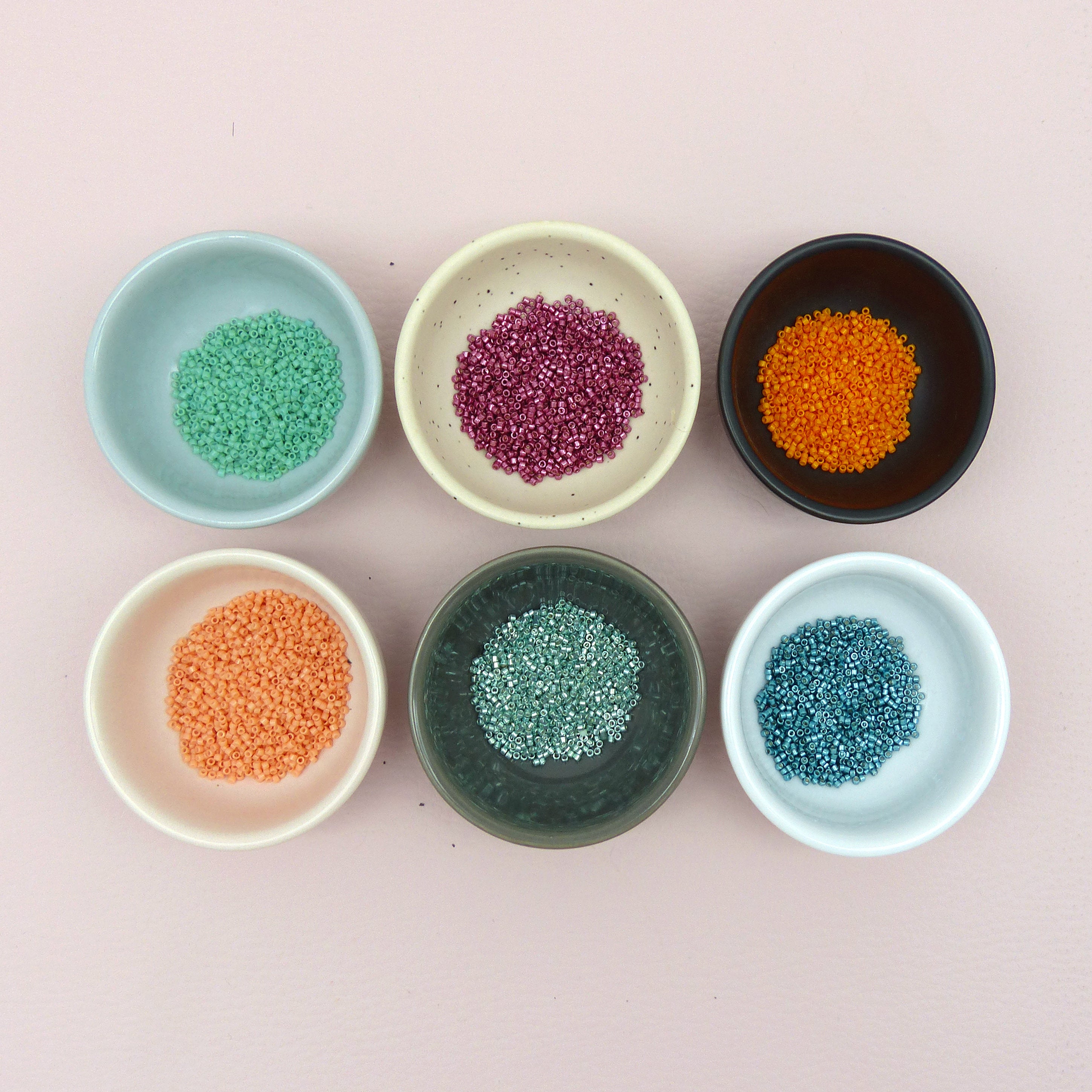 Peut inclure: Six petits bols en céramique remplis de perles minuscules de différentes couleurs. Les couleurs comprennent le vert, le violet, l'orange, le sarcelle et le bleu, sur un fond rose. Les bols sont disposés en deux rangées de trois.