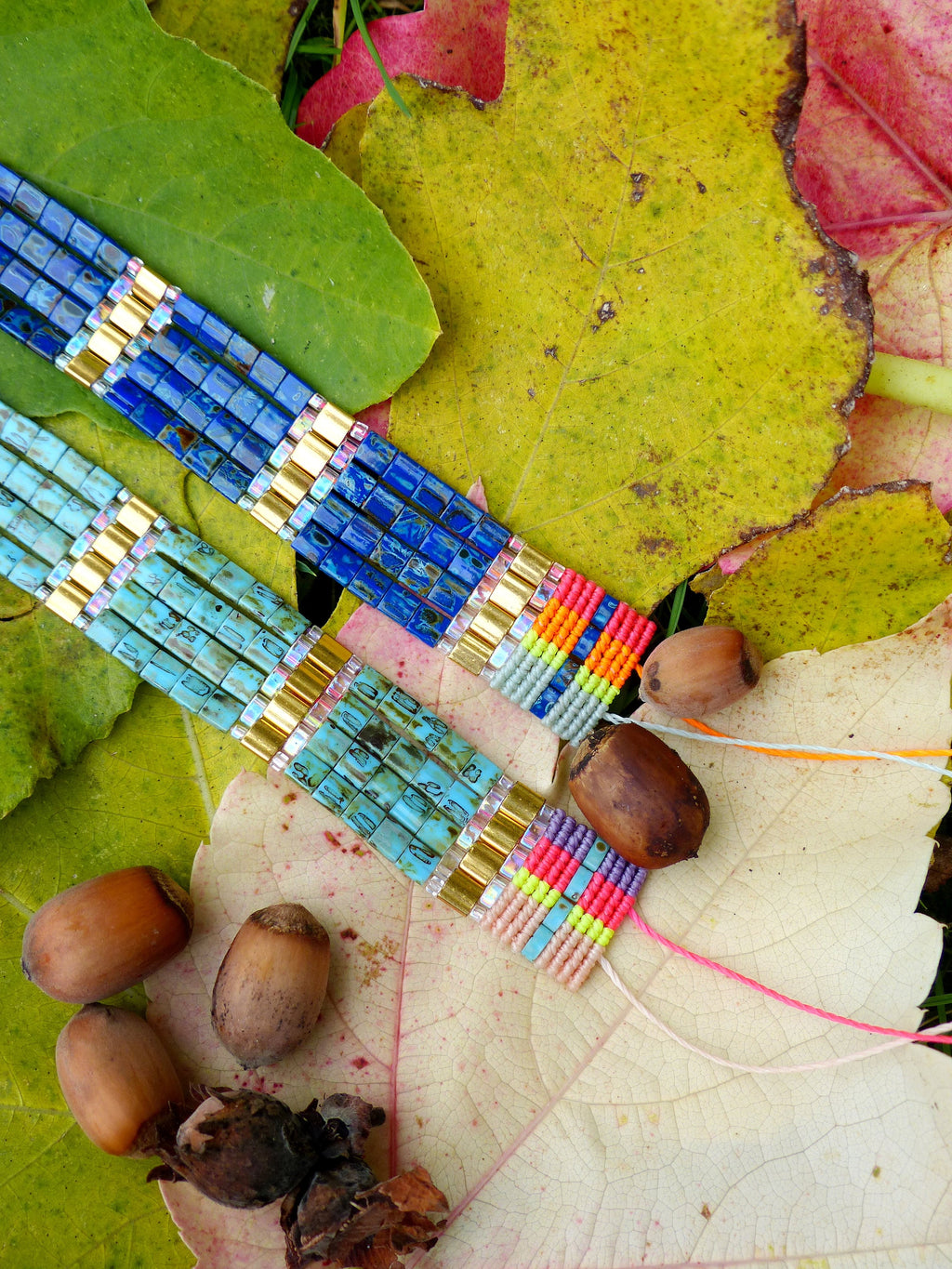 Peut inclure: Deux bracelets en perles colorées avec des accents dorés, posés sur un lit de feuilles d'automne. Les bracelets présentent une variété de couleurs, notamment le bleu, le turquoise, le rose, le jaune et l'orange.