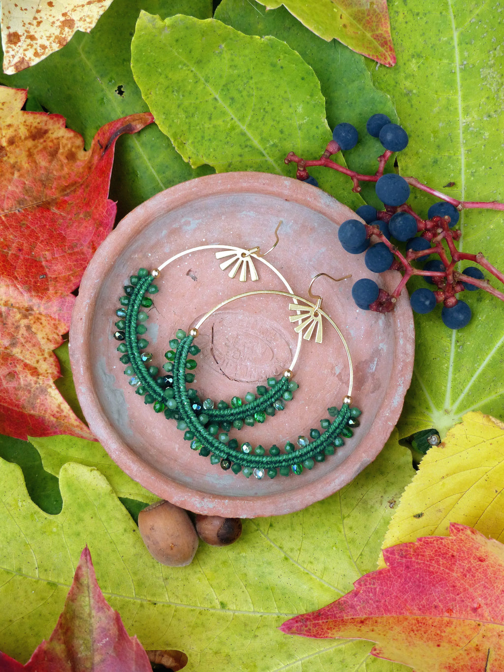 Peut inclure: Une paire de boucles d'oreilles créoles en perles vertes avec des accents dorés. Les boucles d'oreilles sont présentées sur une assiette en terre cuite entourée de feuilles d'automne.