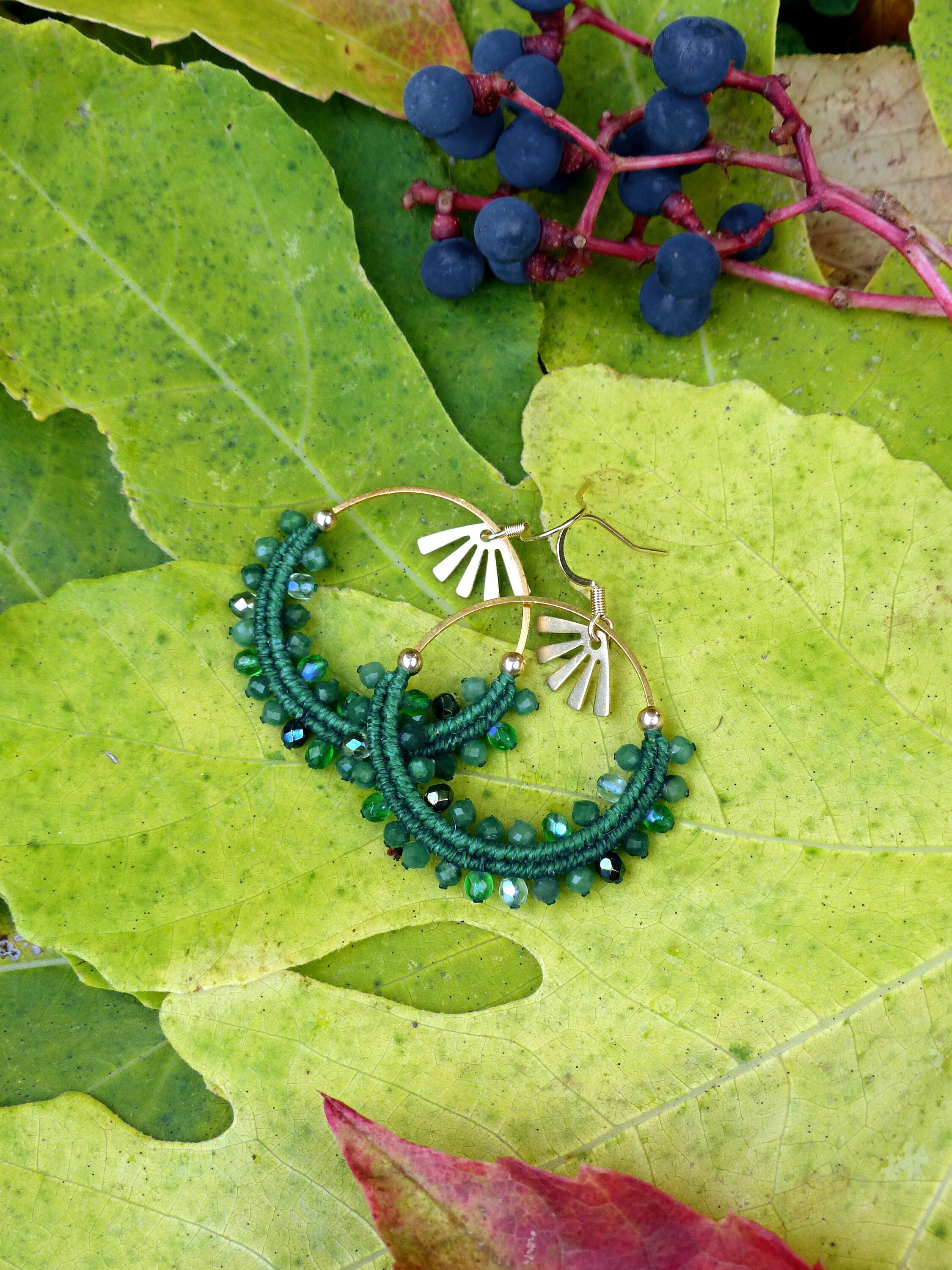 Peut inclure: Boucles d'oreilles créoles dorées avec un motif en forme de soleil et des perles vertes. Les boucles d'oreilles sont posées sur des feuilles vertes et des baies bleu foncé, créant un contraste naturel et vibrant.
