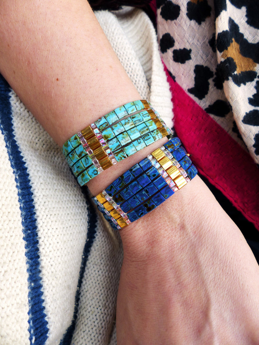 Peut inclure: Deux bracelets superposés sur le poignet d'une personne. Le bracelet du haut est composé de carreaux carrés bleu turquoise et or. Le bracelet du bas est composé de carreaux carrés bleu foncé et or.