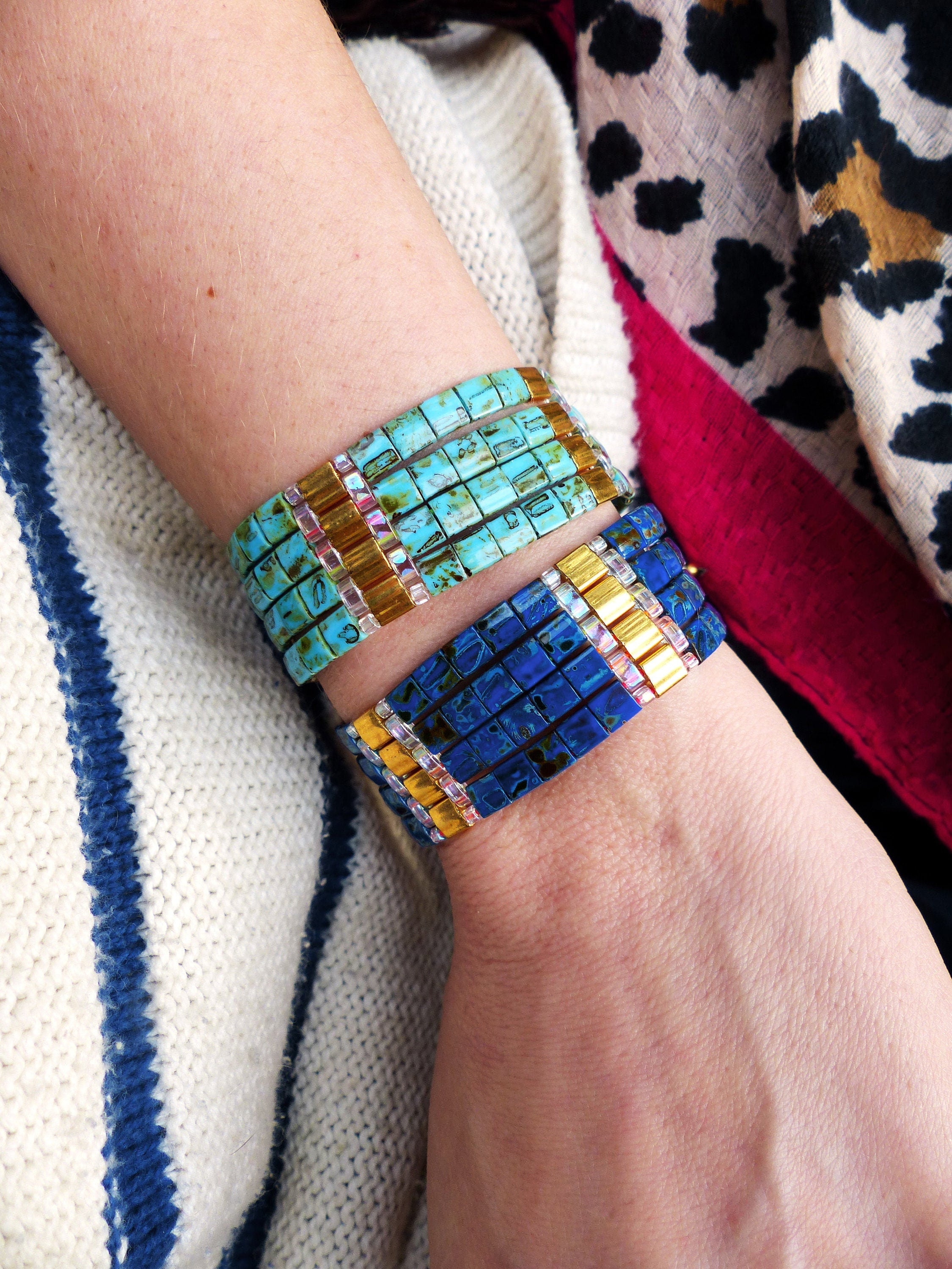 Peut inclure: Deux bracelets superposés sur le poignet d'une personne. Le bracelet du haut est composé de carreaux carrés bleu turquoise et or. Le bracelet du bas est composé de carreaux carrés bleu foncé et or.