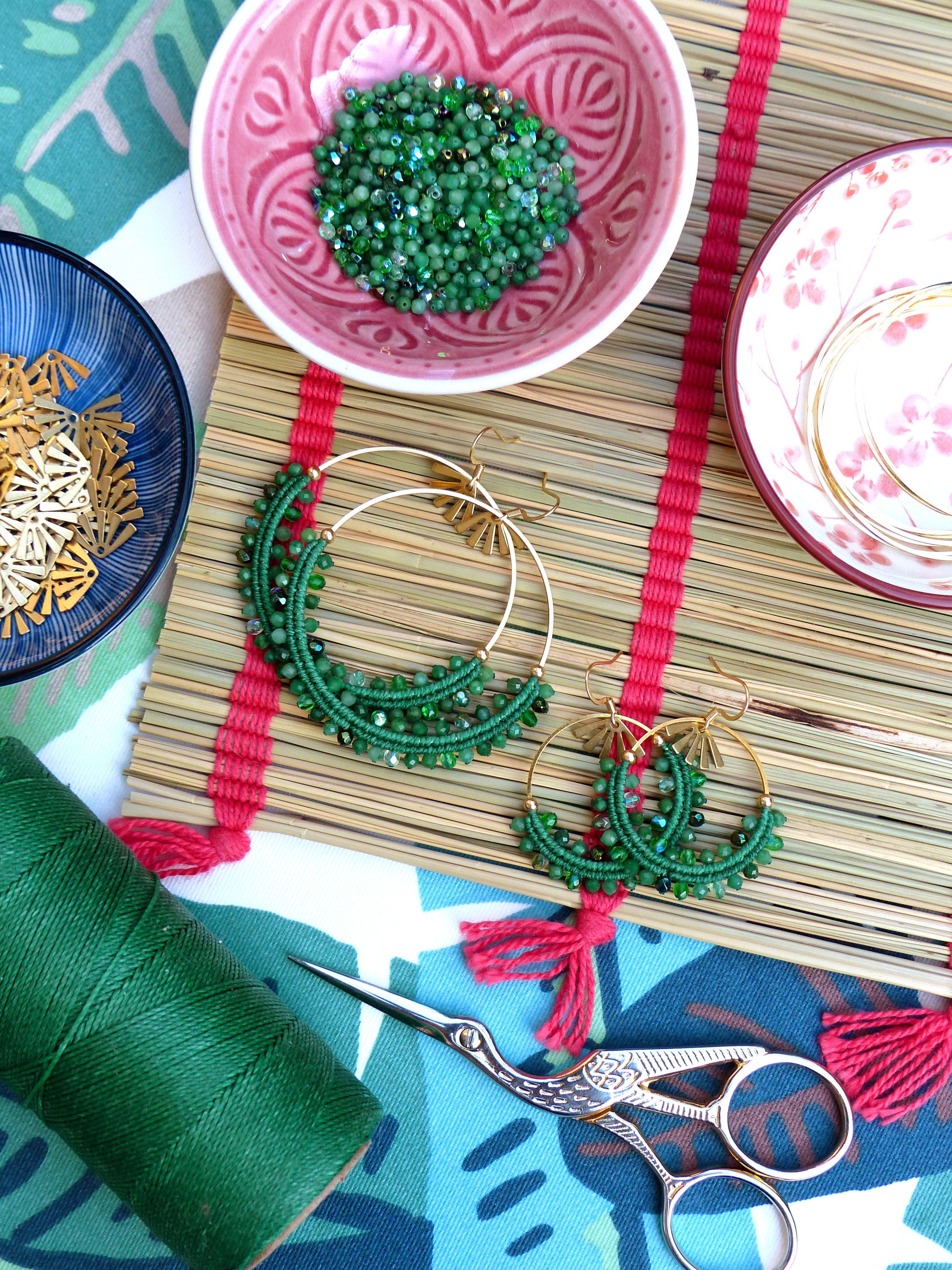 Peut inclure: Une paire de boucles d'oreilles créoles dorées avec des perles vertes, une bobine de fil vert, une paire de ciseaux et un bol de perles vertes sur un tapis en bambou.