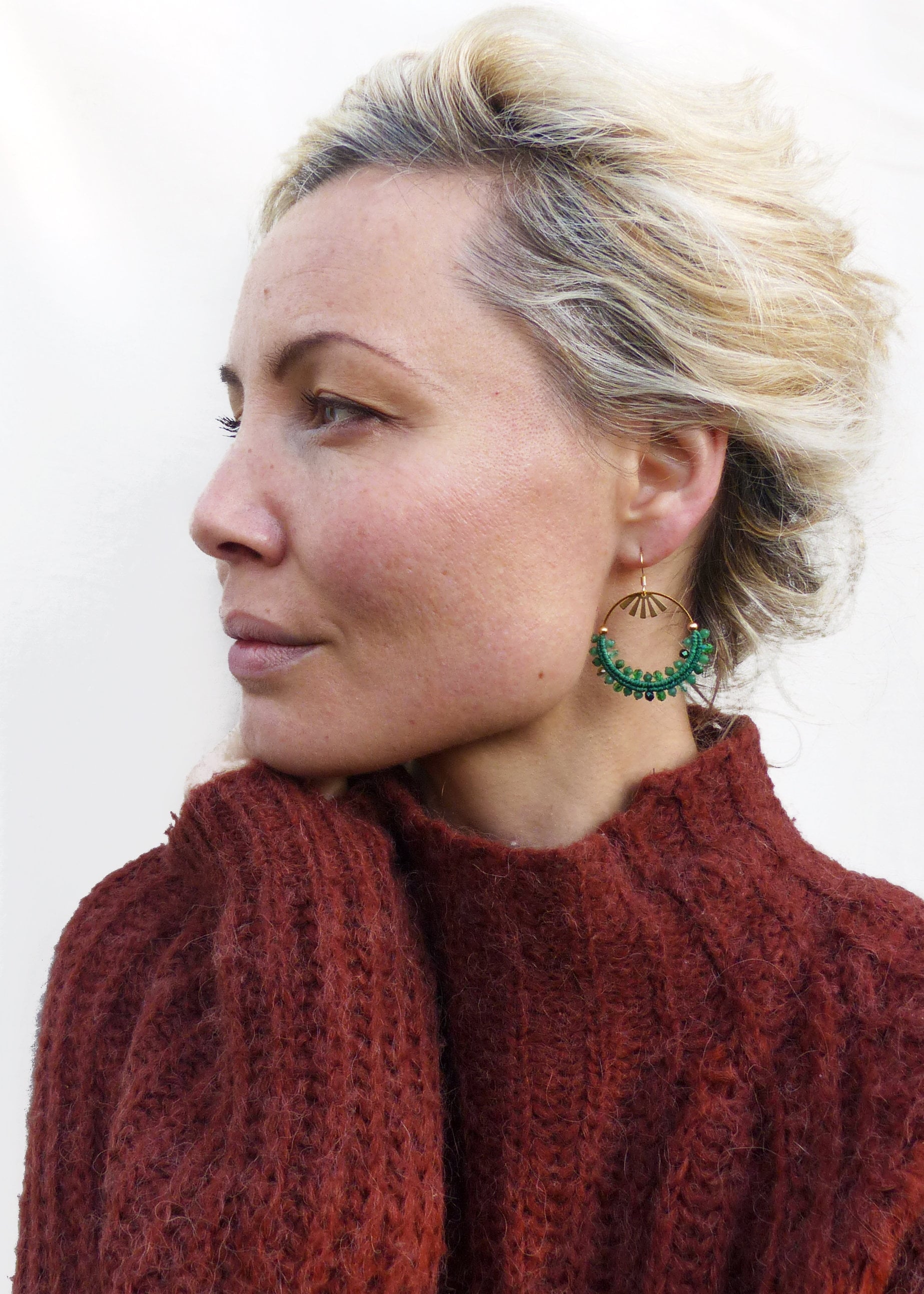 Peut inclure: Une femme porte des boucles d'oreilles dorées en forme de cercle avec un motif de soleil et des accents de perles vertes. Elle porte un pull texturé de couleur rouille. Le fond est un mur blanc uni.