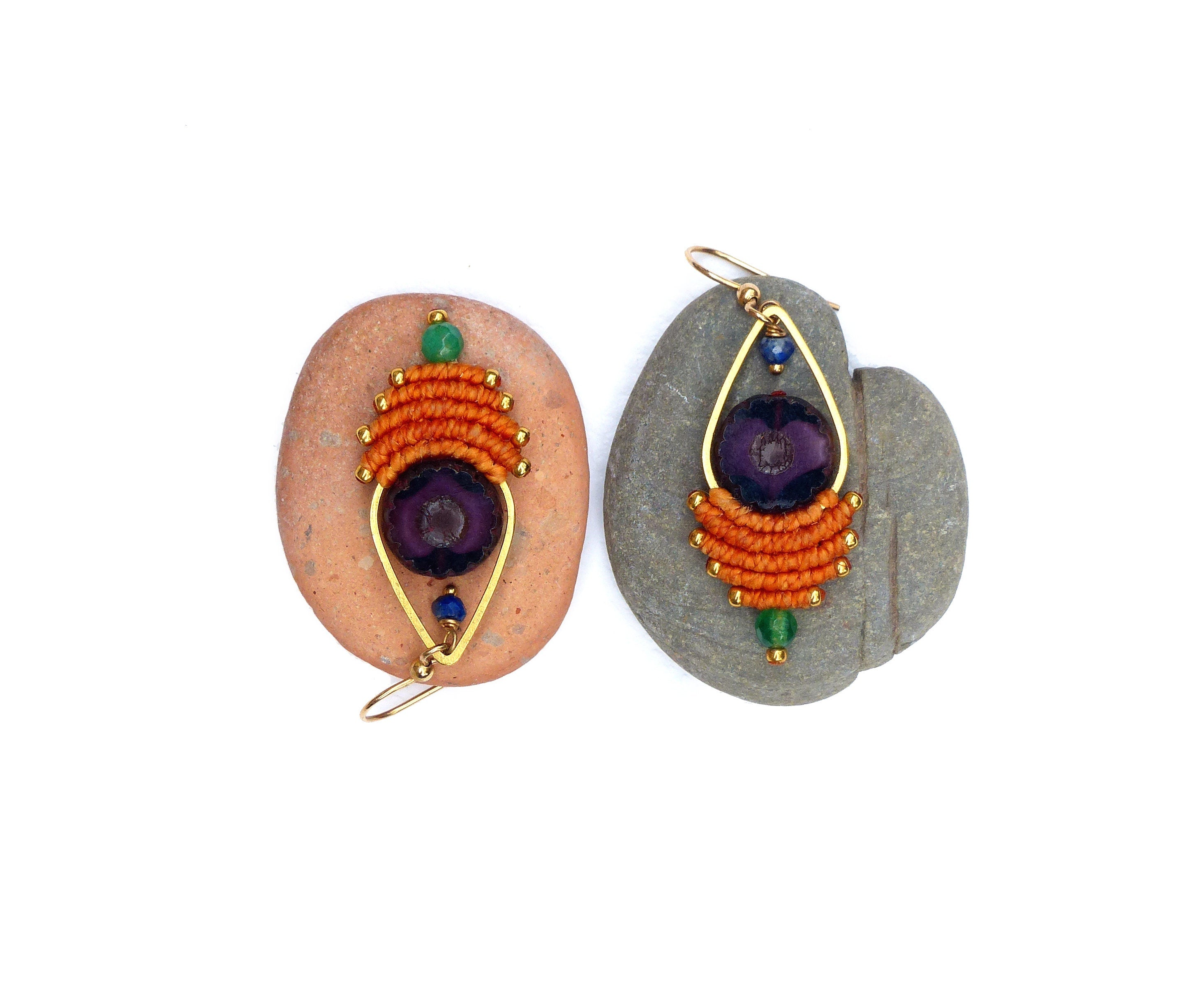 Peut inclure: Une paire de boucles d'oreilles avec une pierre violette, un fil orange et doré, et des perles vertes et bleues. Les boucles d'oreilles sont présentées sur deux pierres grises.