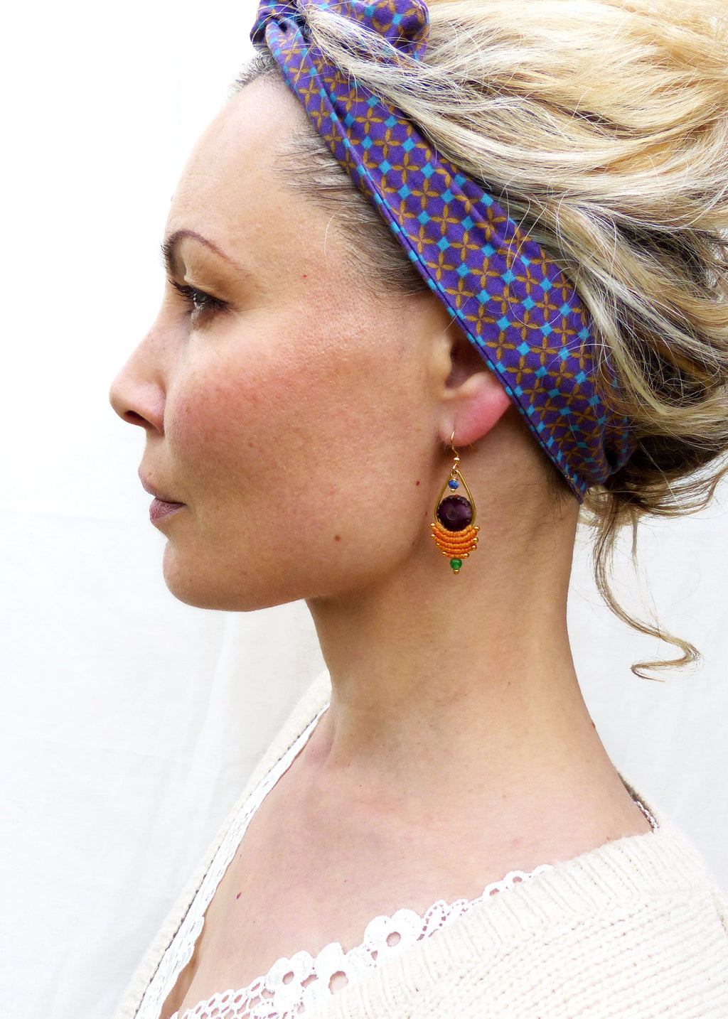 Peut inclure: Une femme portant un bandeau à motif violet et marron avec une paire de boucles d'oreilles pendantes. Les boucles d'oreilles présentent une grosse pierre précieuse violette entourée de perles orange et vertes.