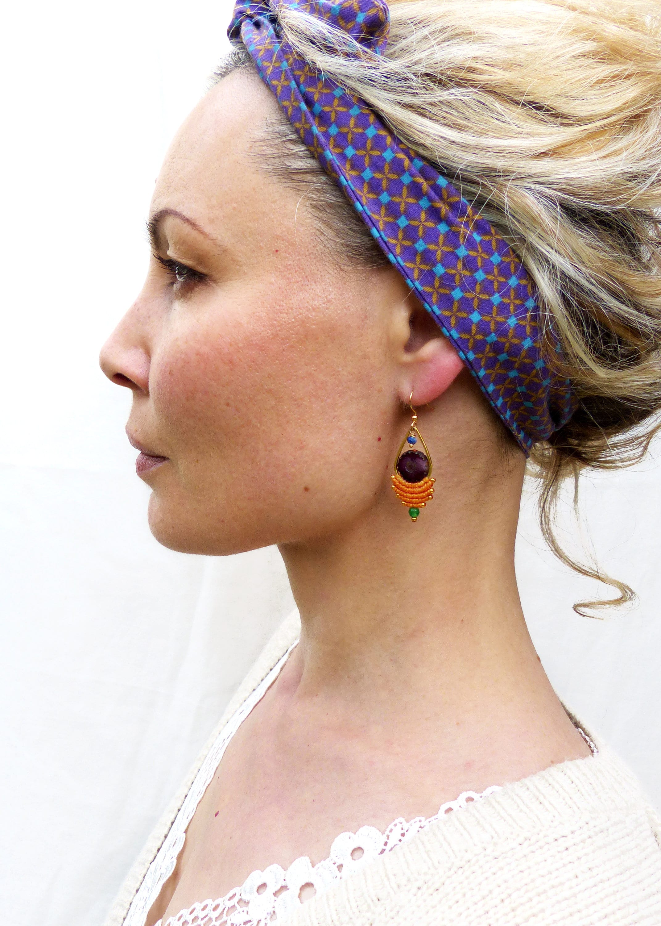 Peut inclure: Une femme portant un bandeau à motif violet et marron avec une paire de boucles d'oreilles pendantes. Les boucles d'oreilles présentent une grosse pierre précieuse violette entourée de perles orange et vertes.