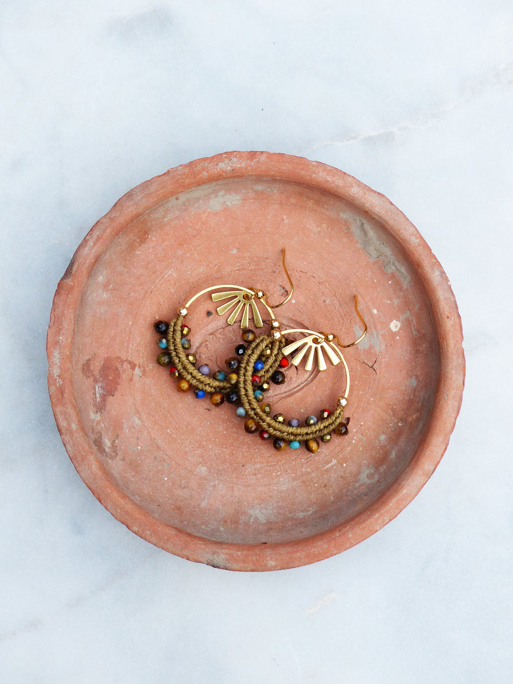 Peut inclure: Une paire de boucles d'oreilles créoles dorées avec un motif soleil et des perles colorées. Les boucles d'oreilles sont présentées sur un plat en terre cuite.