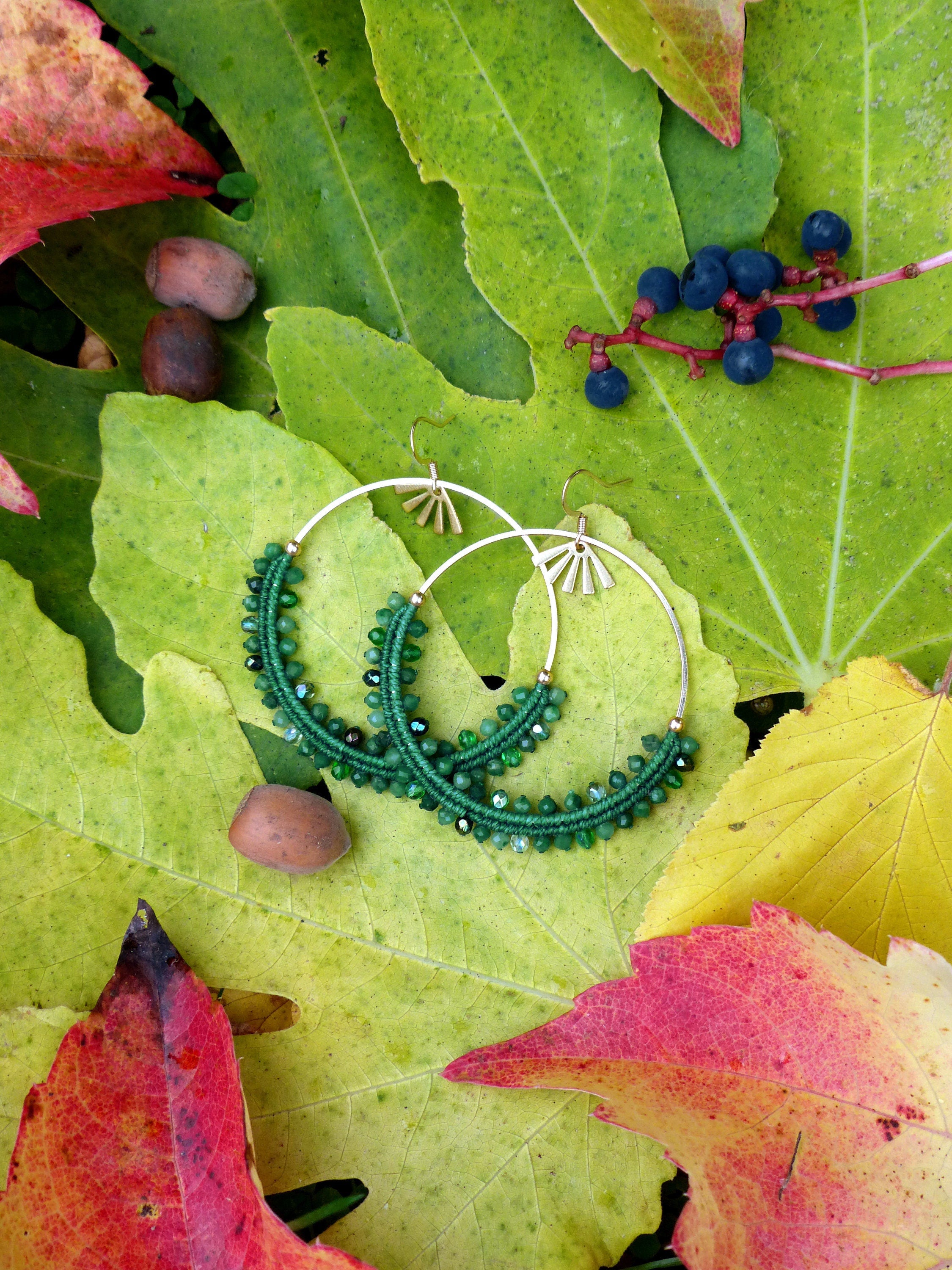 Peut inclure: Une paire de boucles d'oreilles créoles en perles vertes avec des accents dorés. Les boucles d'oreilles sont sur un lit de feuilles vertes et jaunes.