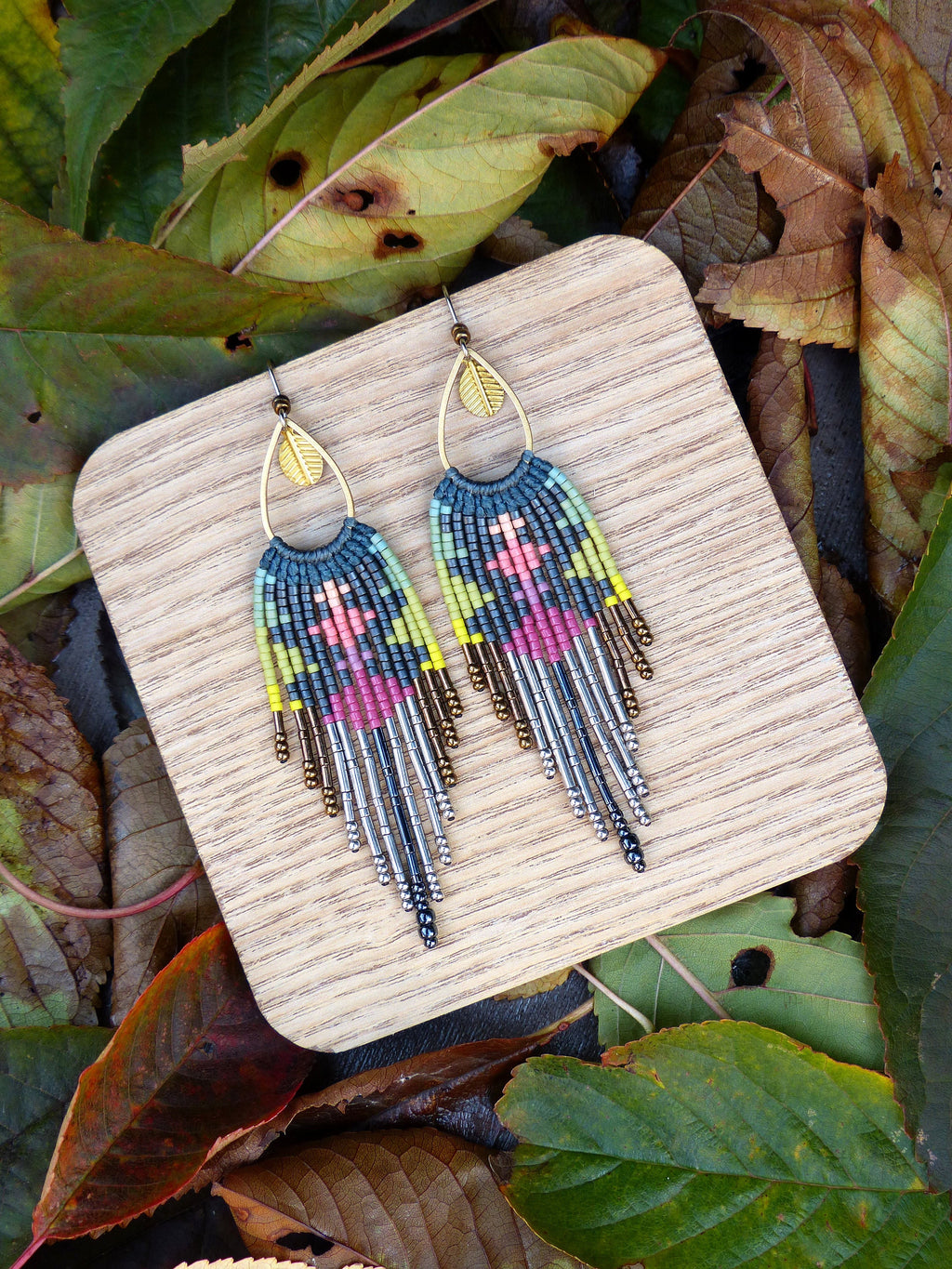 Peut inclure: Une paire de boucles d'oreilles en perles avec un charme en feuille d'or. Les boucles d'oreilles sont faites avec une variété de couleurs, y compris le rose, le bleu, le vert et le jaune. Les boucles d'oreilles sont suspendues à un présentoir en bois.