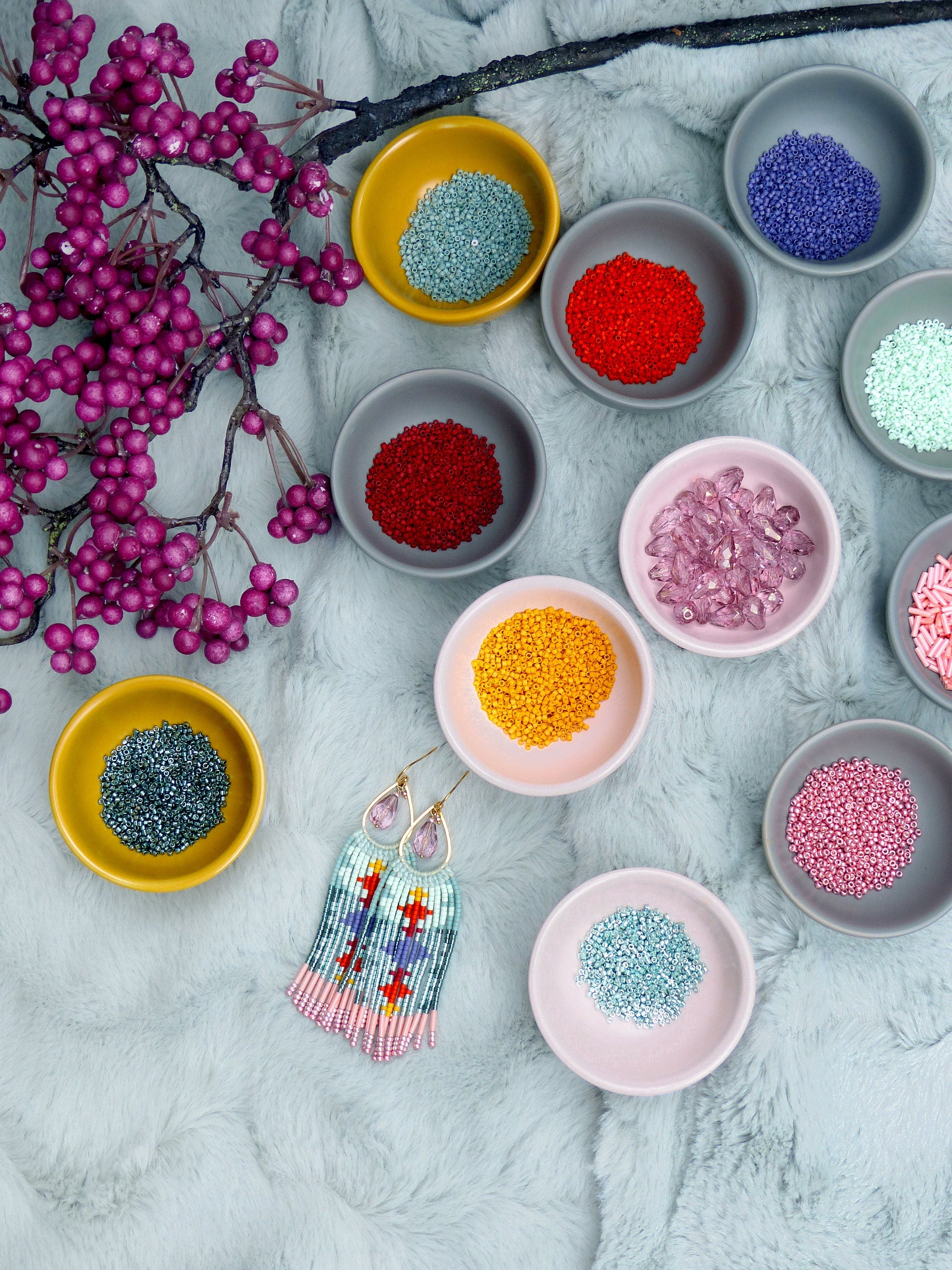Peut inclure: Vue aérienne de perles colorées dans de petits bols, à côté d'une paire de boucles d'oreilles perlées. Les boucles d'oreilles présentent un motif bleu clair, rose et rouge. Les bols contiennent des perles de différentes couleurs, dont le rouge, le jaune et le bleu. Une branche avec des baies violettes est également présente.