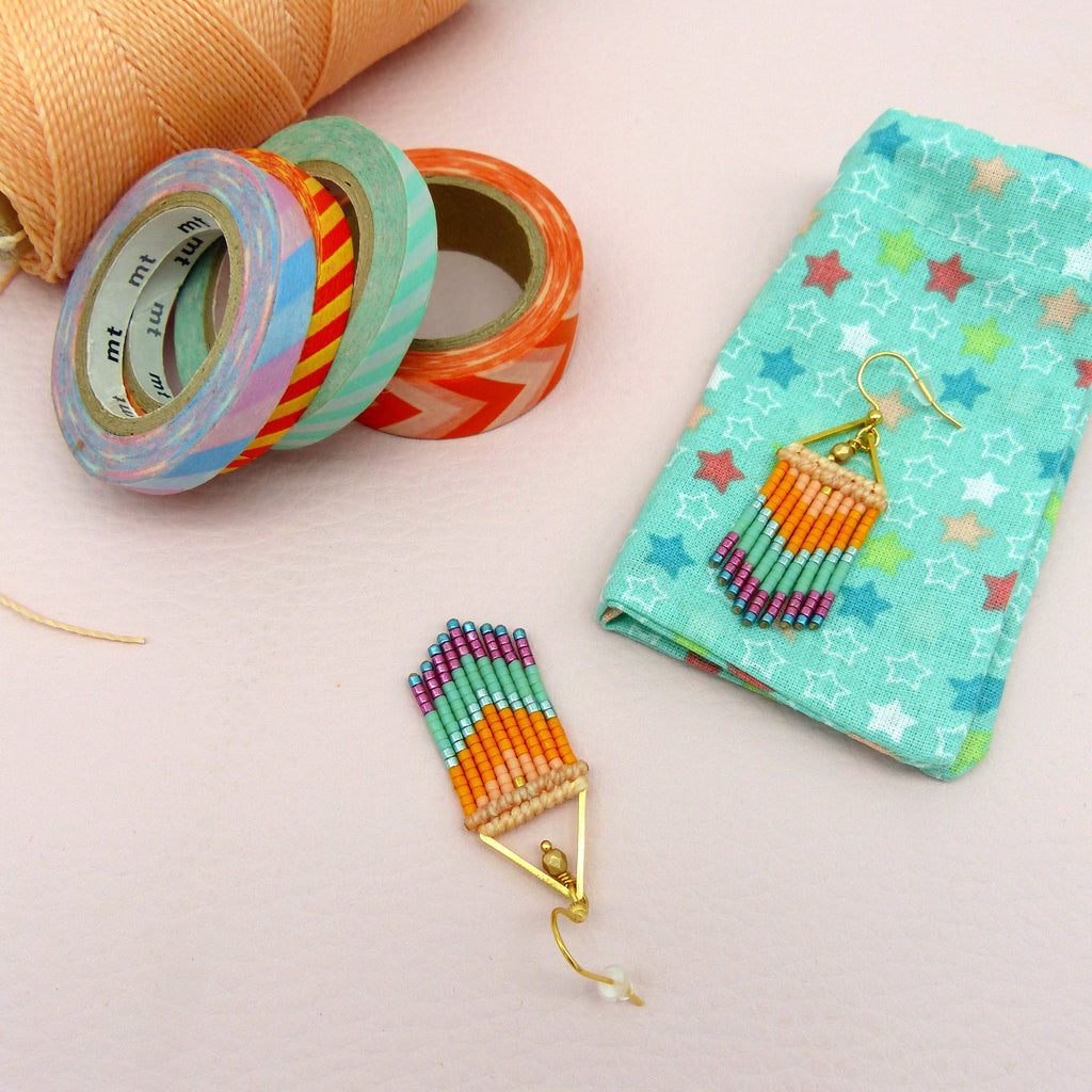 Peut inclure: Boucles d'oreilles faites à la main avec un cadre triangulaire doré, ornées de perles colorées orange, rose et turquoise. Un petit pochon turquoise à motifs d'étoiles et des rouleaux de ruban adhésif décoratif sont également présents.