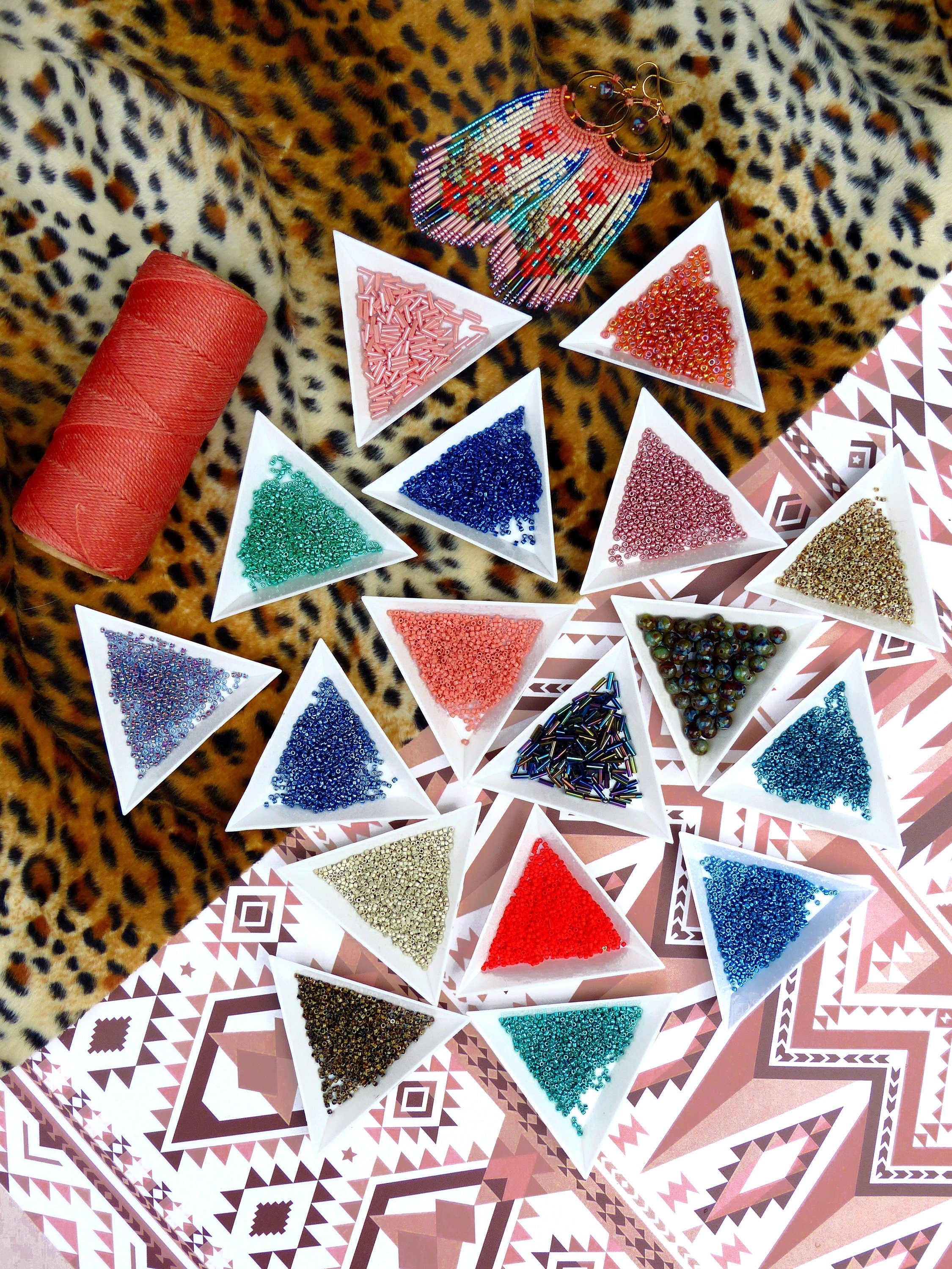 Peut inclure: Assortiment de perles de rocaille colorées dans de petits plats triangulaires sur une surface à motifs. Les perles sont disposées dans différentes couleurs, notamment le rouge, le bleu, le vert, l'or et l'argent. Il y a également une bobine de fil rouge dans l'image.