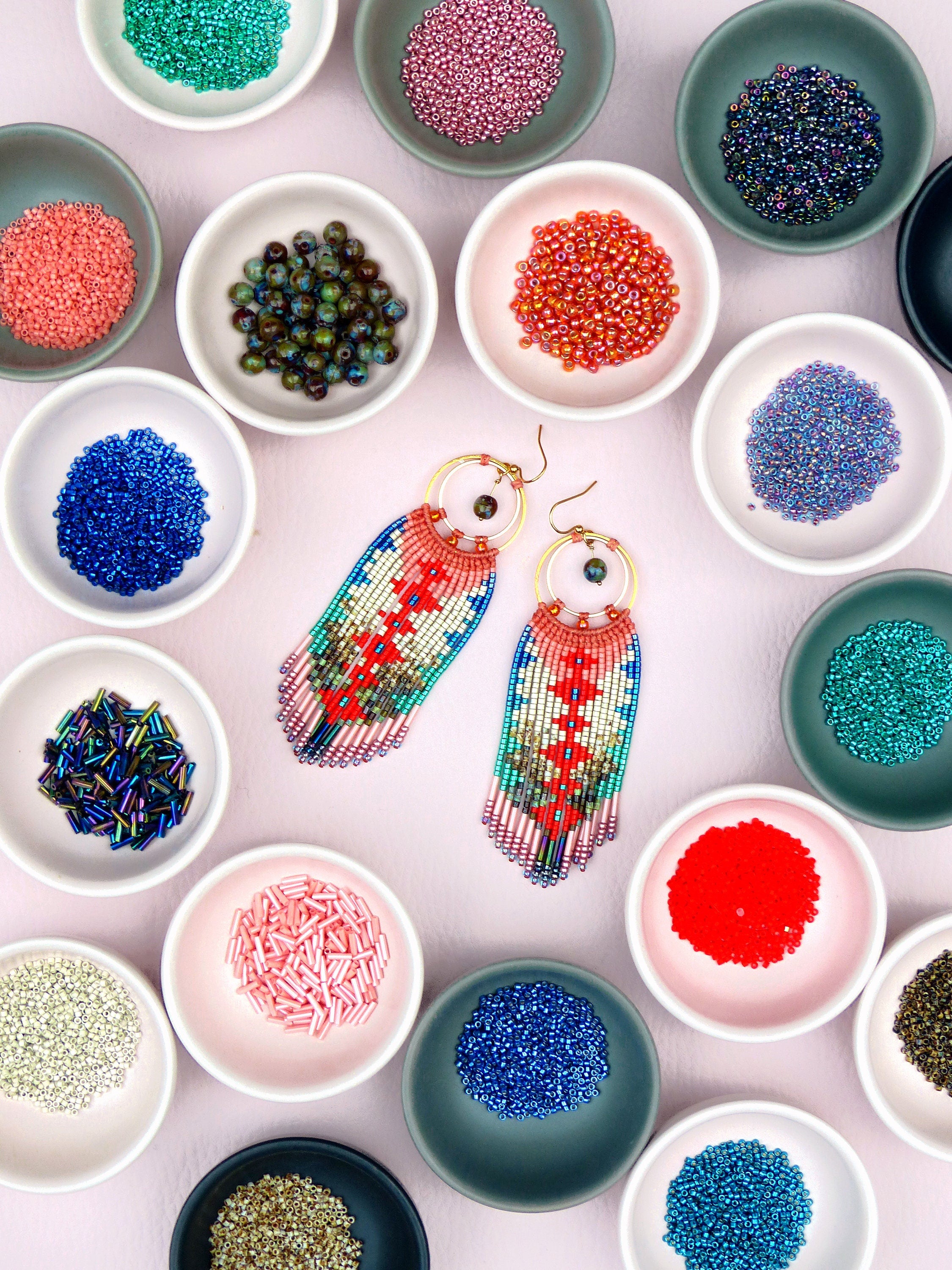 Peut inclure: Une paire de boucles d'oreilles en perles colorées avec un motif géométrique. Les boucles d'oreilles sont présentées sur une surface rose avec de petits bols de perles de différentes couleurs.