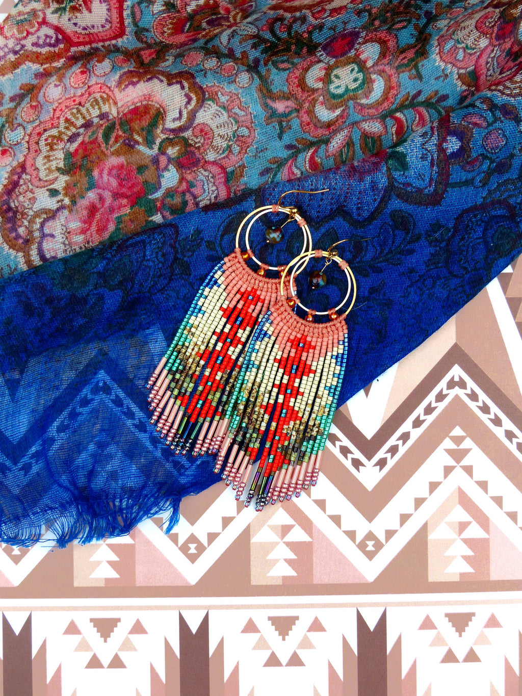 Peut inclure: Une paire de longues boucles d'oreilles en perles avec un design en frange. Les boucles d'oreilles sont faites de perles rouges, bleues, vertes et dorées. Les boucles d'oreilles sont suspendues à un tissu à motifs bleus et roses.