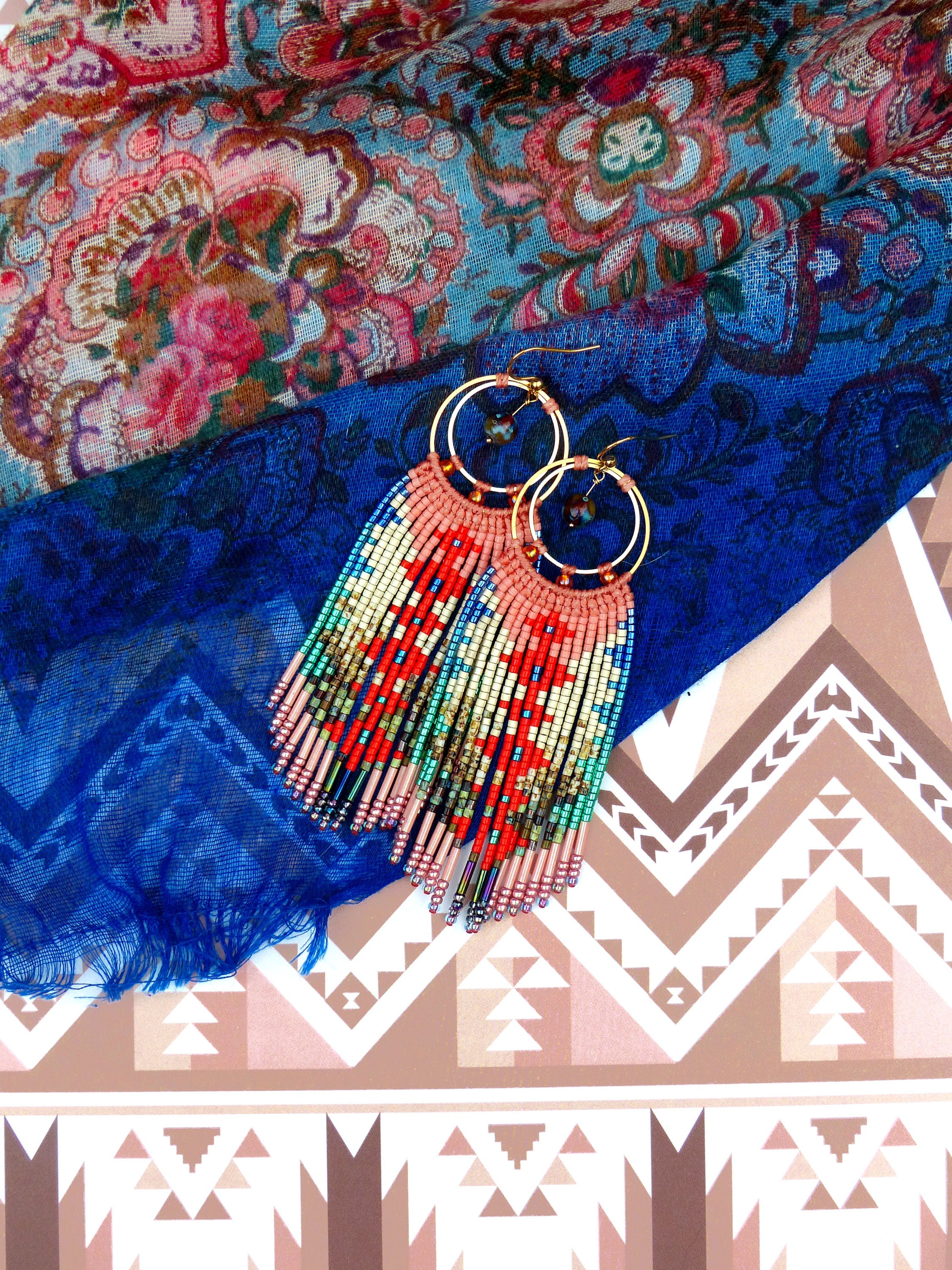 Peut inclure: Une paire de longues boucles d'oreilles en perles avec un design en frange. Les boucles d'oreilles sont faites de perles rouges, bleues, vertes et dorées. Les boucles d'oreilles sont suspendues à un tissu à motifs bleus et roses.