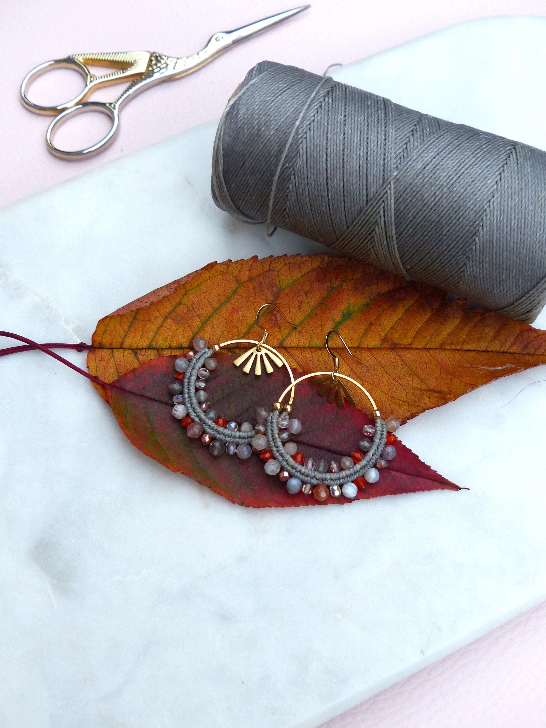 Peut inclure: Une paire de boucles d'oreilles créoles faites à la main avec des perles grises et orange et des accents dorés. Les boucles d'oreilles sont présentées sur une surface blanche avec une bobine de fil gris et une paire de ciseaux.