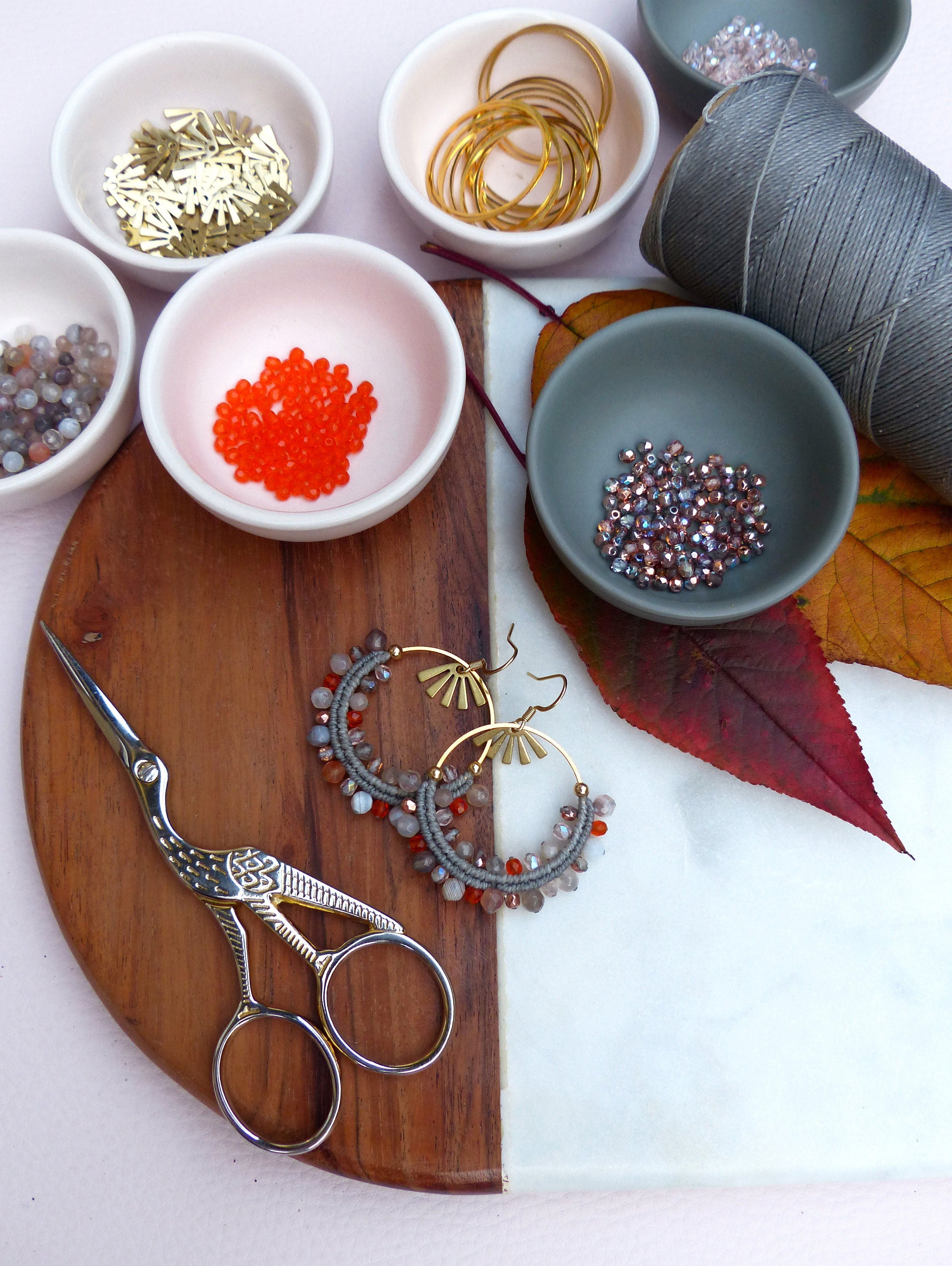 Peut inclure: Une paire de boucles d'oreilles faites à la main avec des perles grises et orange, des cercles dorés et un charme en forme d'éventail doré. Les boucles d'oreilles sont présentées sur une planche à découper blanche et brune avec diverses perles et fournitures.