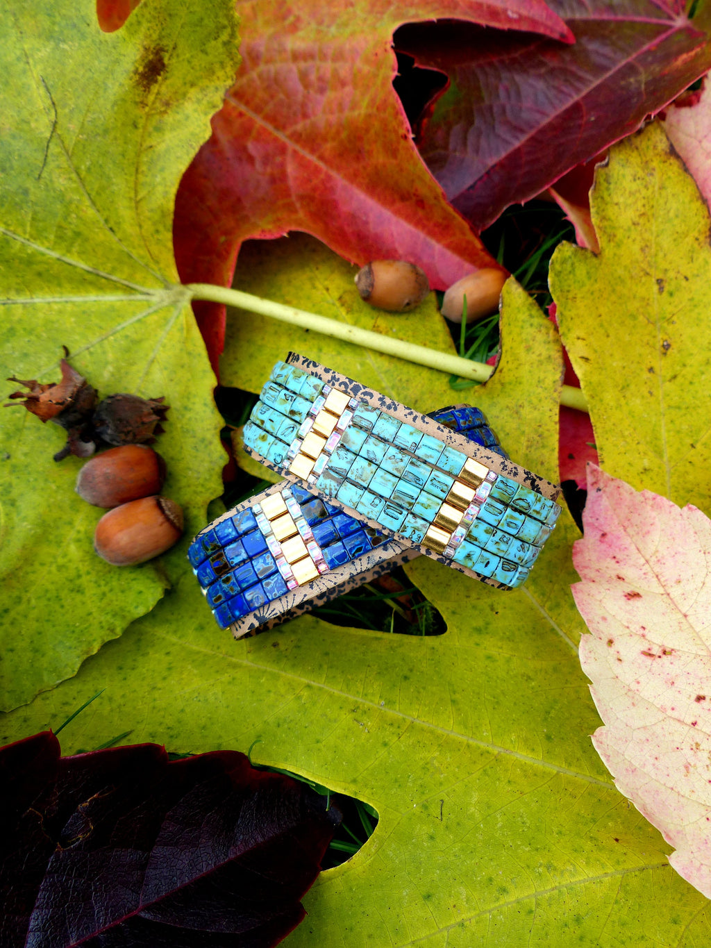 Peut inclure: Deux bracelets en mosaïque bleu et or avec un dos en liège, posés sur un lit de feuilles d'automne.