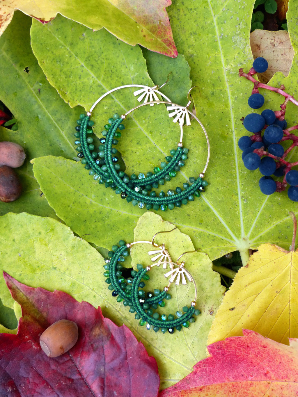 Peut inclure: Boucles d'oreilles créoles dorées avec un motif en forme de soleil et des perles vertes. Les boucles d'oreilles sont posées sur un fond de feuilles d'automne colorées, vertes, rouges et jaunes.