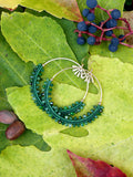 Peut inclure: Une paire de boucles d'oreilles créoles vertes et dorées avec un délicat motif perlé. Les boucles d'oreilles sont photographiées sur un lit de feuilles vertes.