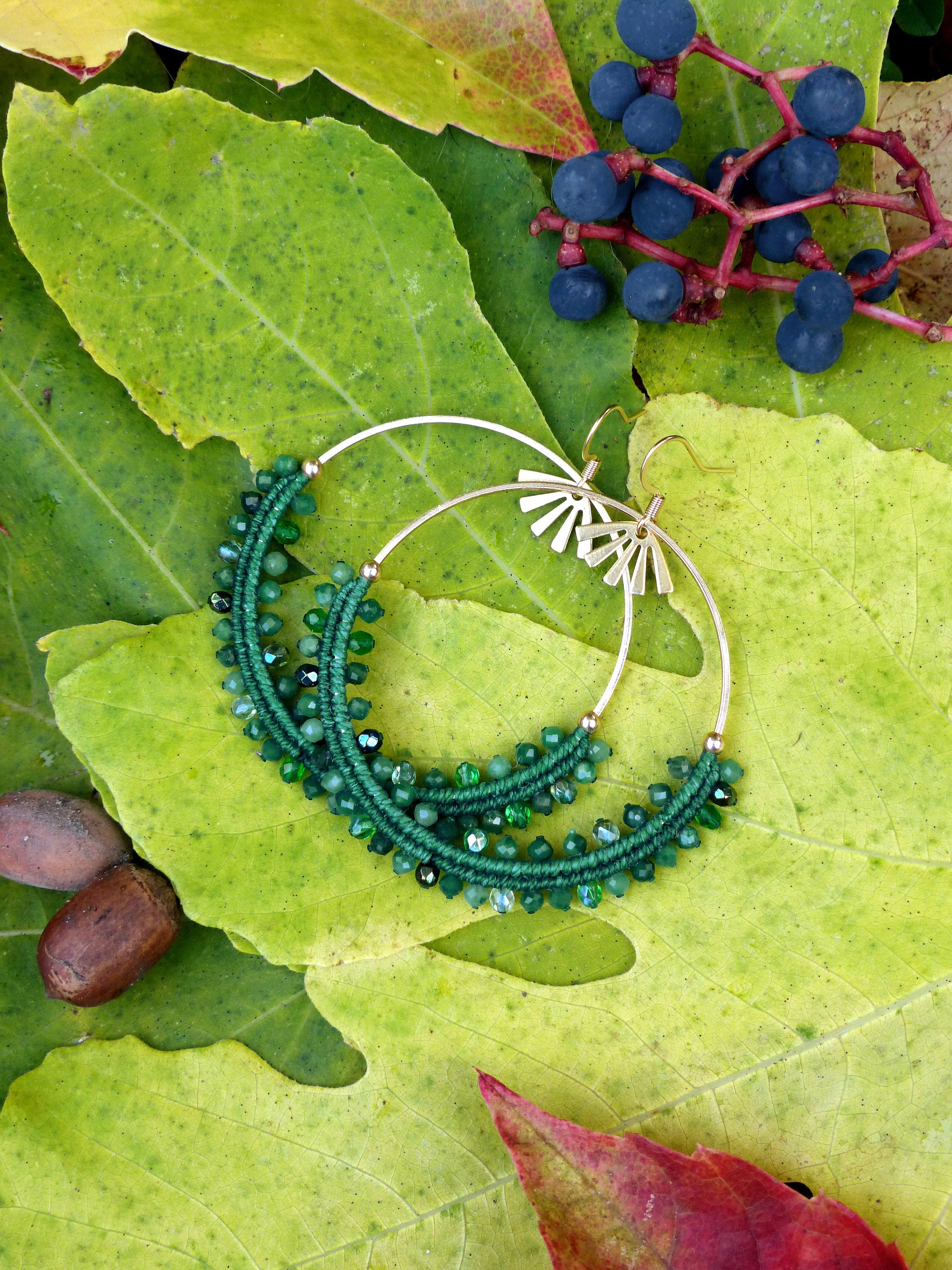Peut inclure: Une paire de boucles d'oreilles créoles vertes et dorées avec un délicat motif perlé. Les boucles d'oreilles sont photographiées sur un lit de feuilles vertes.
