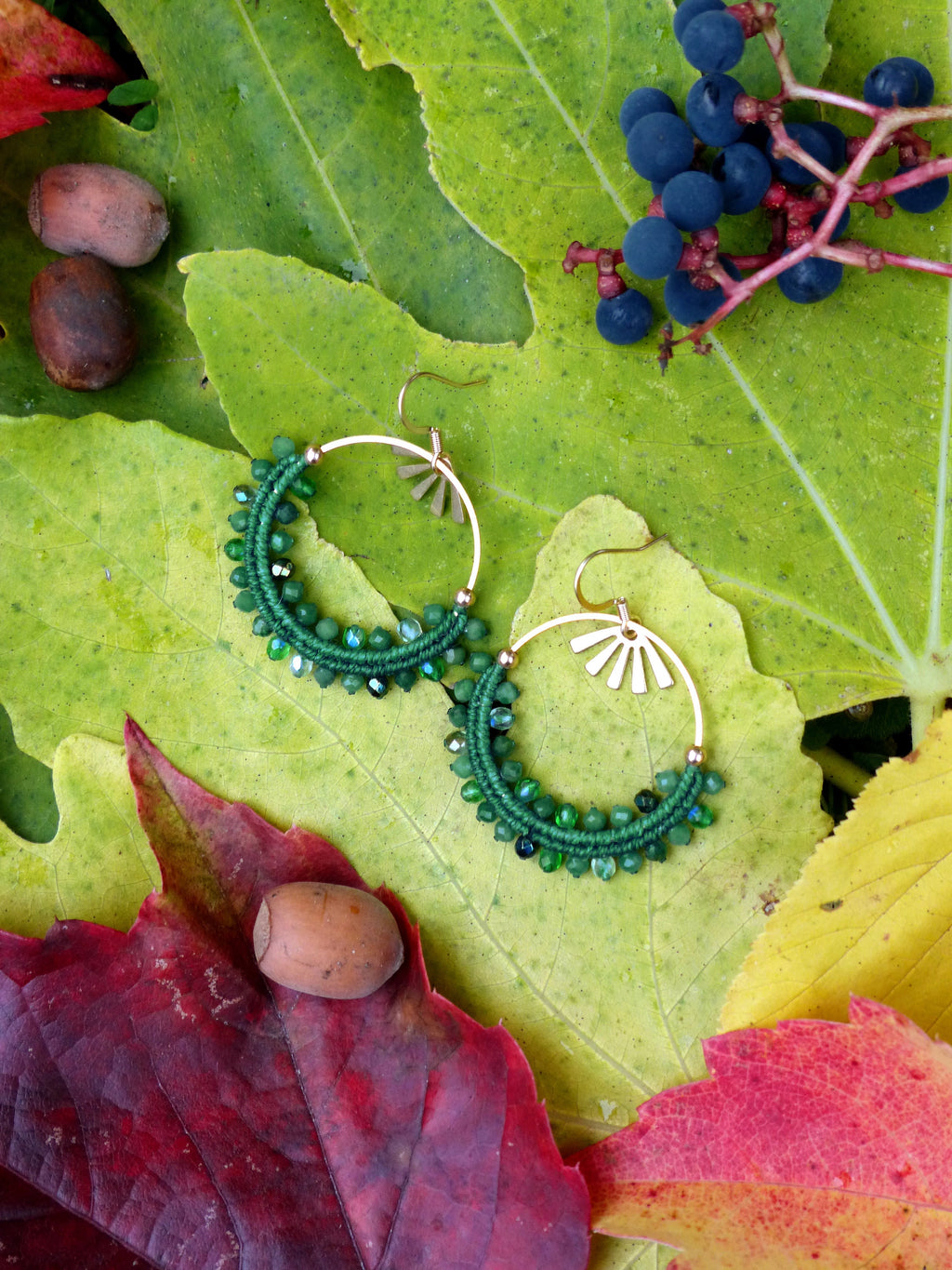 Peut inclure: Boucles d'oreilles créoles dorées avec des détails de perles vertes. Les boucles d'oreilles sont ornées de petites perles vertes rondes et d'un motif de soleil. Présentées sur un lit de feuilles d'automne colorées.