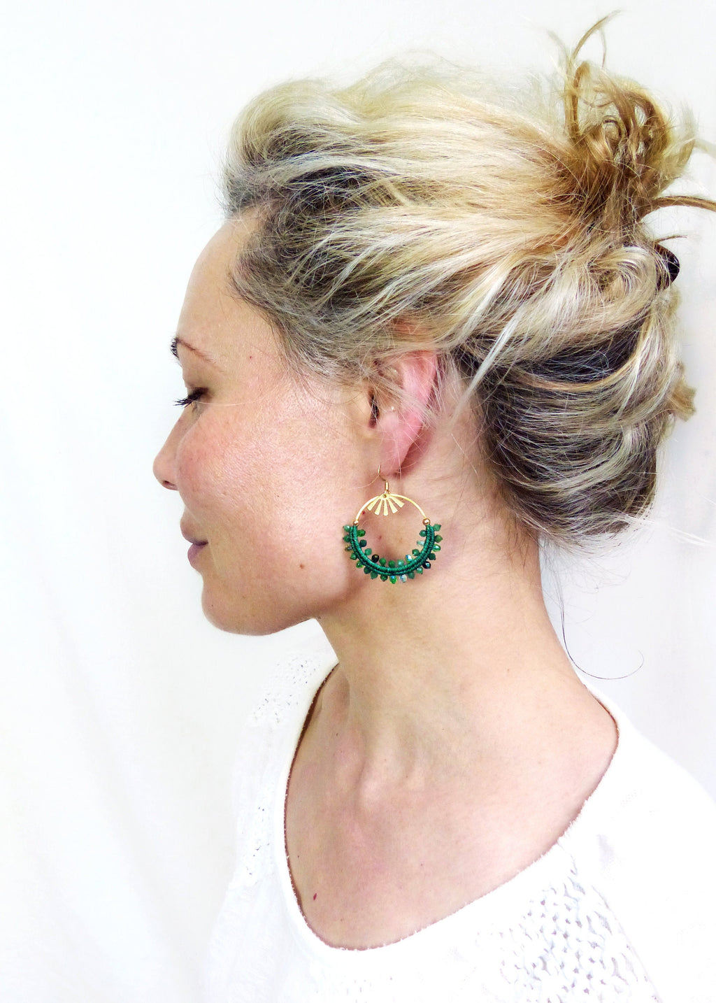 Peut inclure: Profil d'une personne portant de grandes boucles d'oreilles vertes. Les boucles d'oreilles ont une partie supérieure dorée avec un motif de soleil et une section inférieure circulaire verte perlée. La personne a les cheveux blonds attachés en chignon.