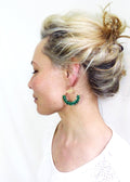 Peut inclure: Profil d'une personne portant de grandes boucles d'oreilles vertes. Les boucles d'oreilles ont une partie supérieure dorée avec un motif de soleil et une section inférieure circulaire verte perlée. La personne a les cheveux blonds attachés en chignon.