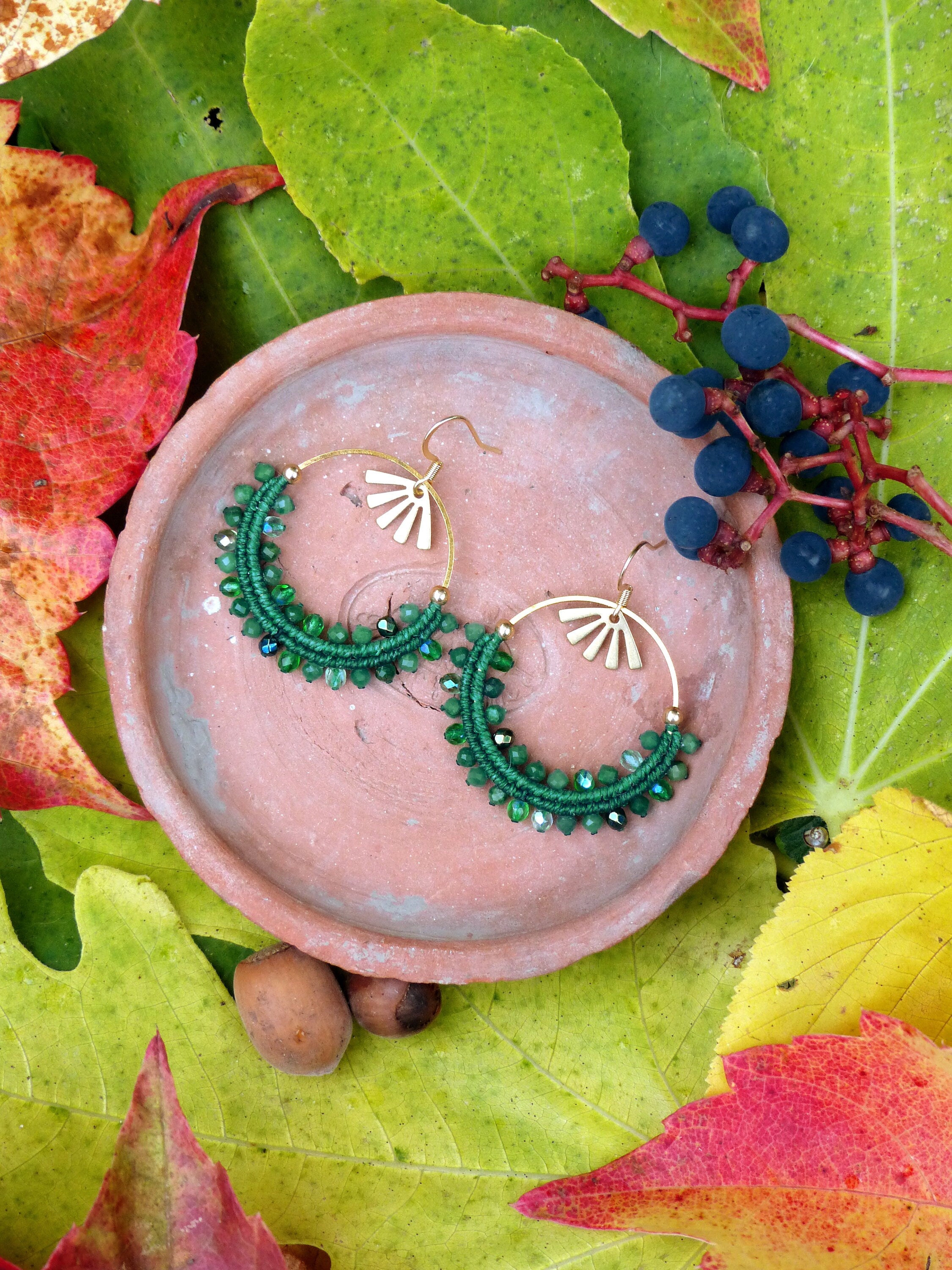 Peut inclure: Une paire de boucles d'oreilles créoles vertes faites à la main, ornées de perles et de breloques dorées en forme de soleil. Les boucles d'oreilles sont présentées sur une petite coupelle ronde de couleur terre cuite, entourée de feuilles d'automne colorées et de baies.