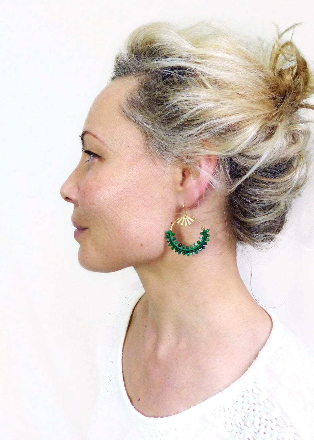 Peut inclure: Vue de profil d'une personne portant des boucles d'oreilles créoles vertes perlées avec des détails dorés. Les boucles d'oreilles ont une forme de demi-cercle avec un dessus en forme d'éventail. La personne a les cheveux blonds attachés.