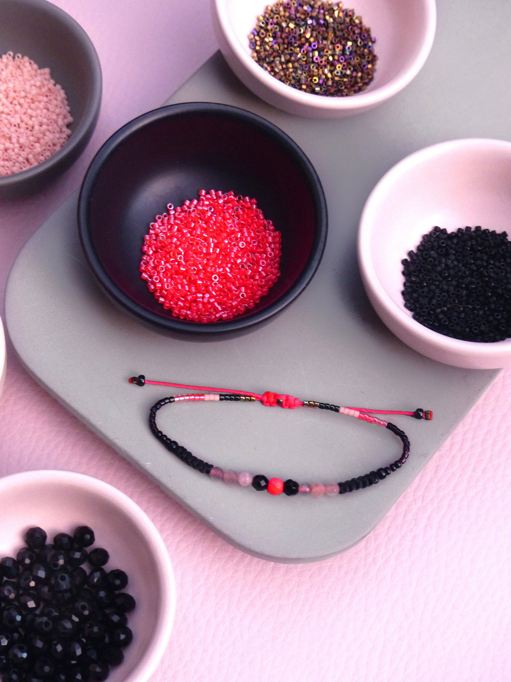Peut inclure: Gros plan sur un bracelet composé de perles noires, roses et rouges. Le bracelet est posé sur une surface grise avec de petits bols de perles de différentes couleurs.