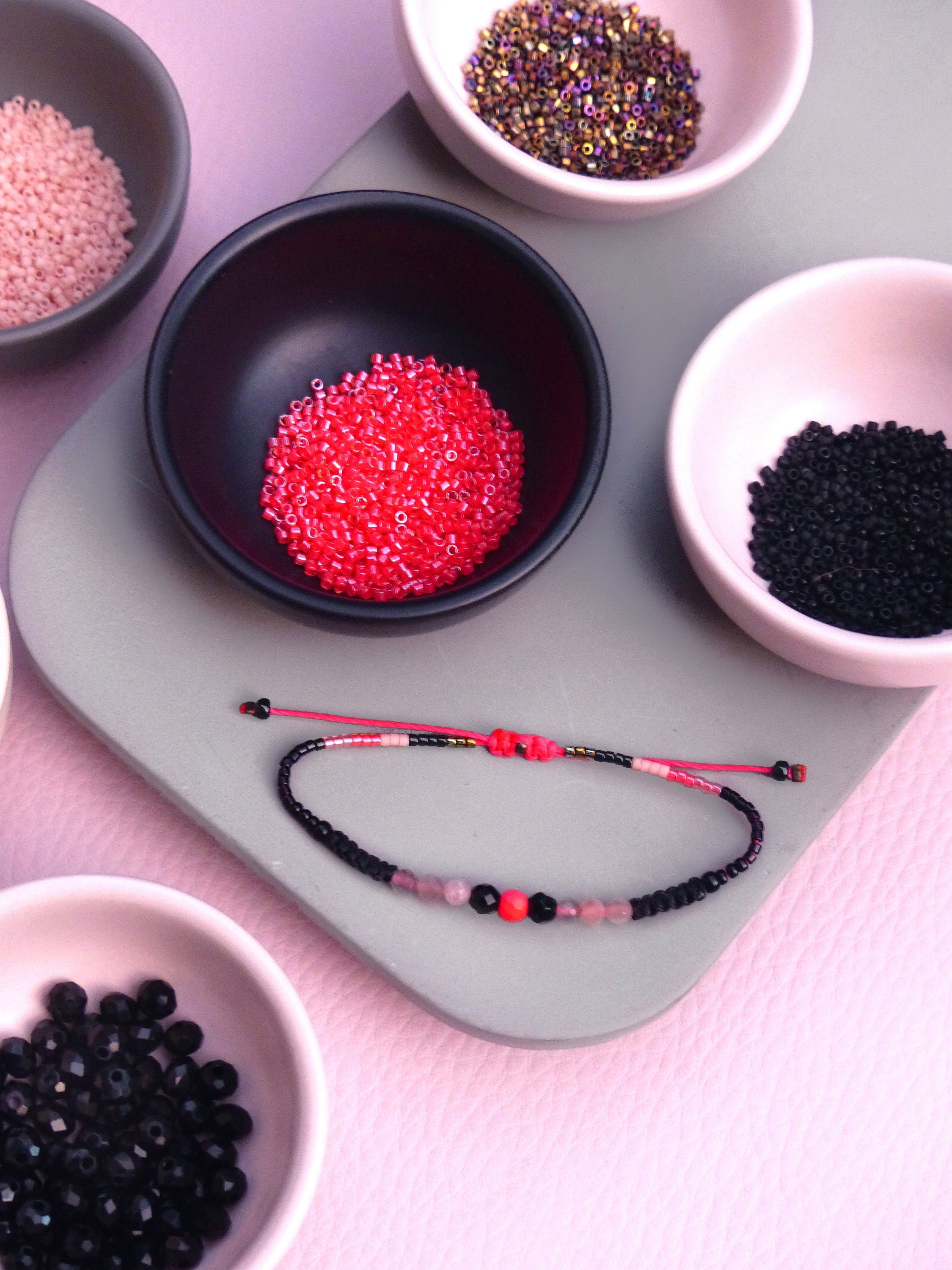 Peut inclure: Gros plan sur un bracelet composé de perles noires, roses et rouges. Le bracelet est posé sur une surface grise avec de petits bols de perles de différentes couleurs.