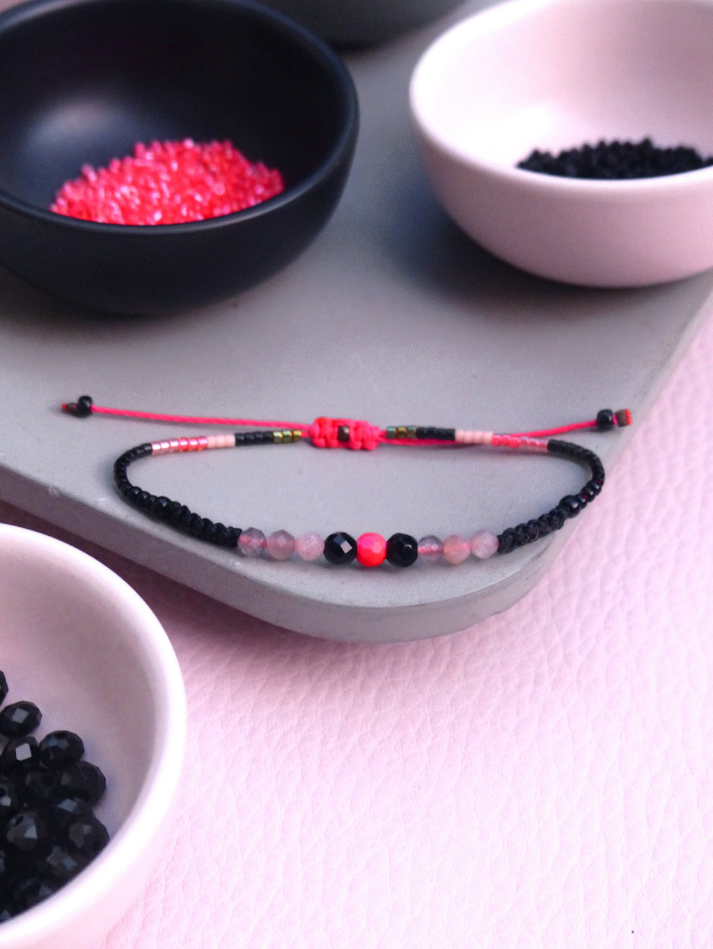 Peut inclure: Un bracelet en perles rose et noir avec une perle rose et noire au centre. Le bracelet est sur une surface grise avec de petits bols de perles en arrière-plan.