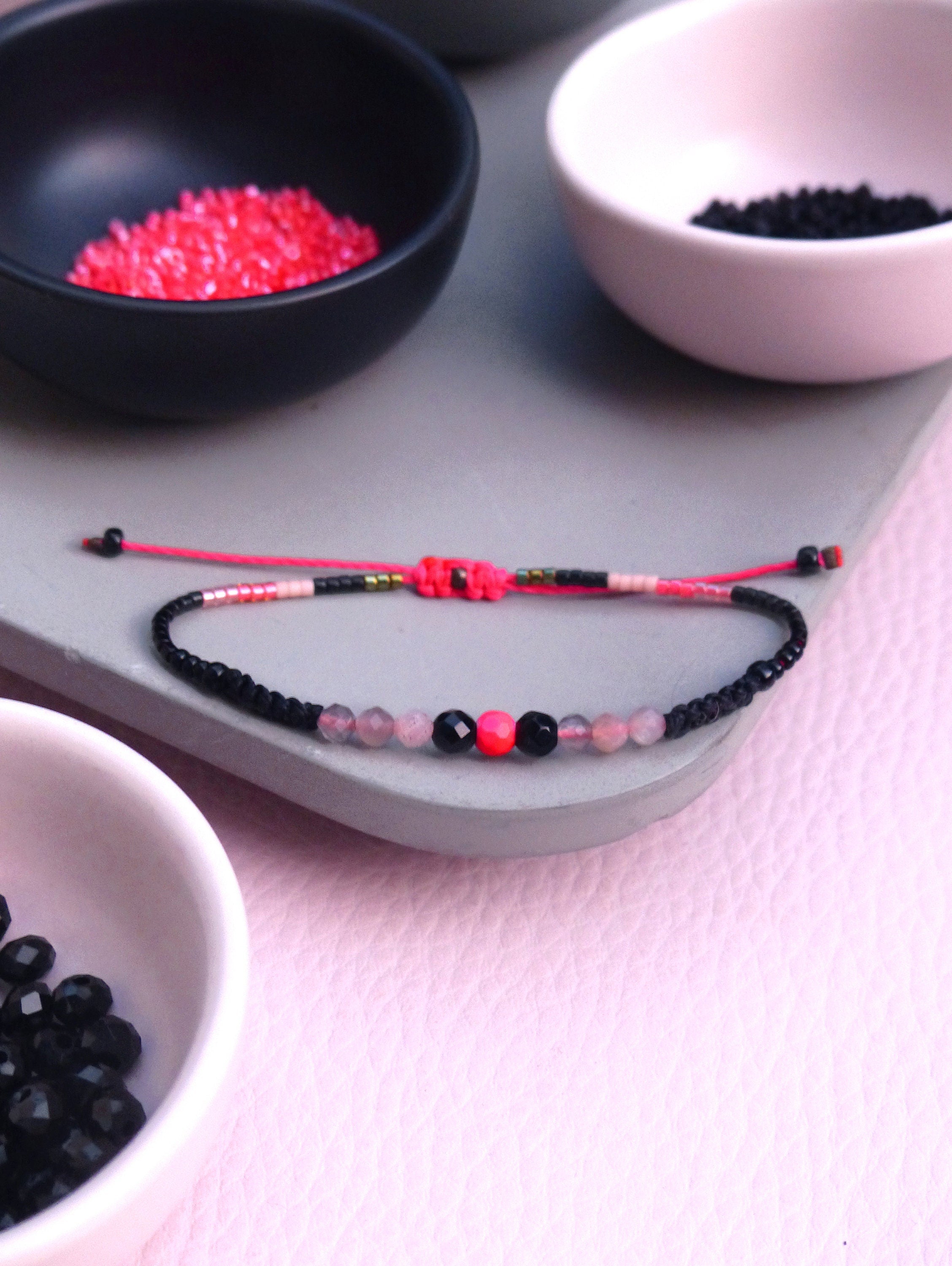 Peut inclure: Un bracelet en perles rose et noir avec une perle rose et noire au centre. Le bracelet est sur une surface grise avec de petits bols de perles en arrière-plan.