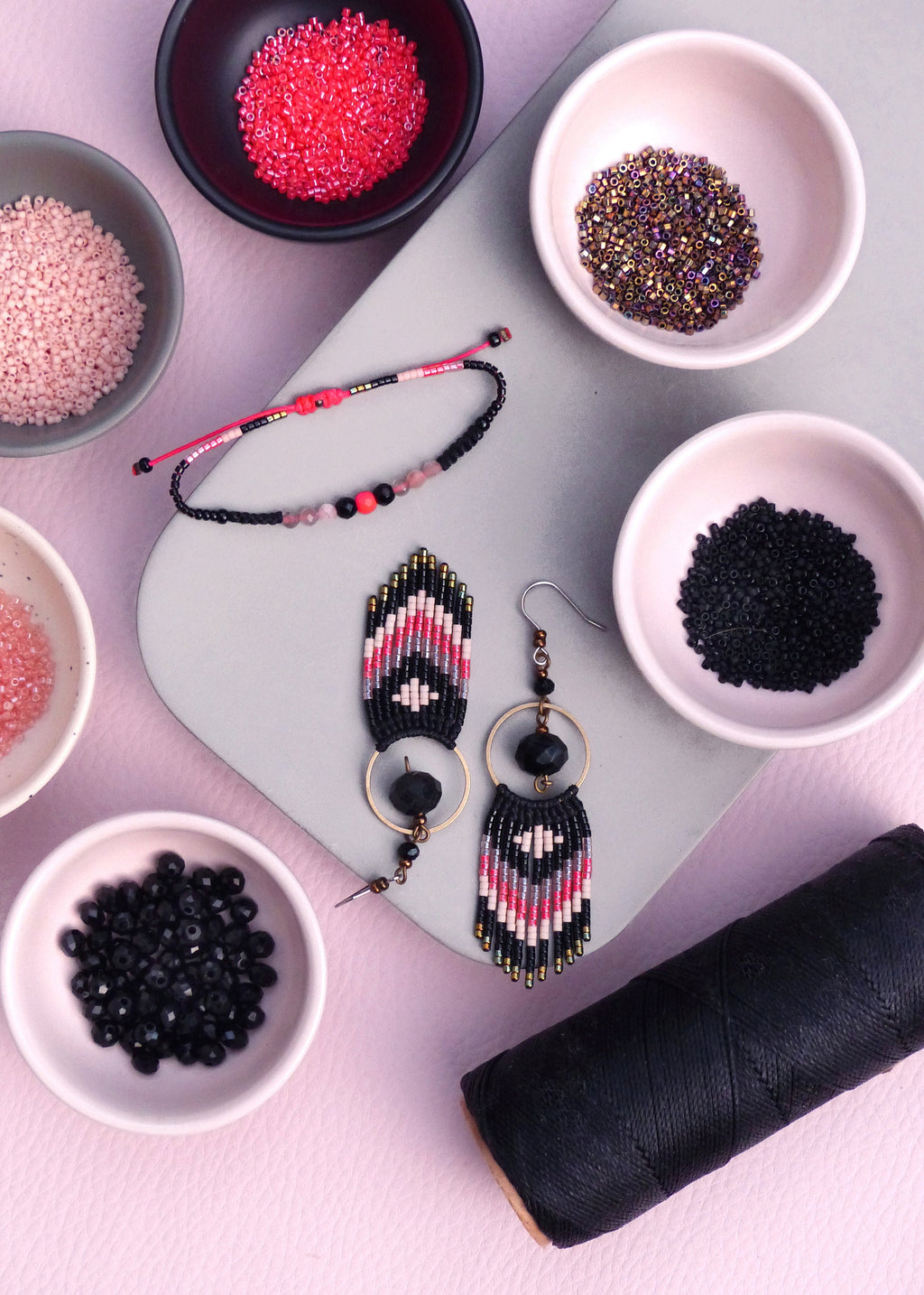 Peut inclure: Une vue de dessus de fournitures de fabrication de bijoux, y compris des perles noires, roses et rouges, du fil noir et deux paires de boucles d'oreilles en perles avec des motifs géométriques noirs et roses.