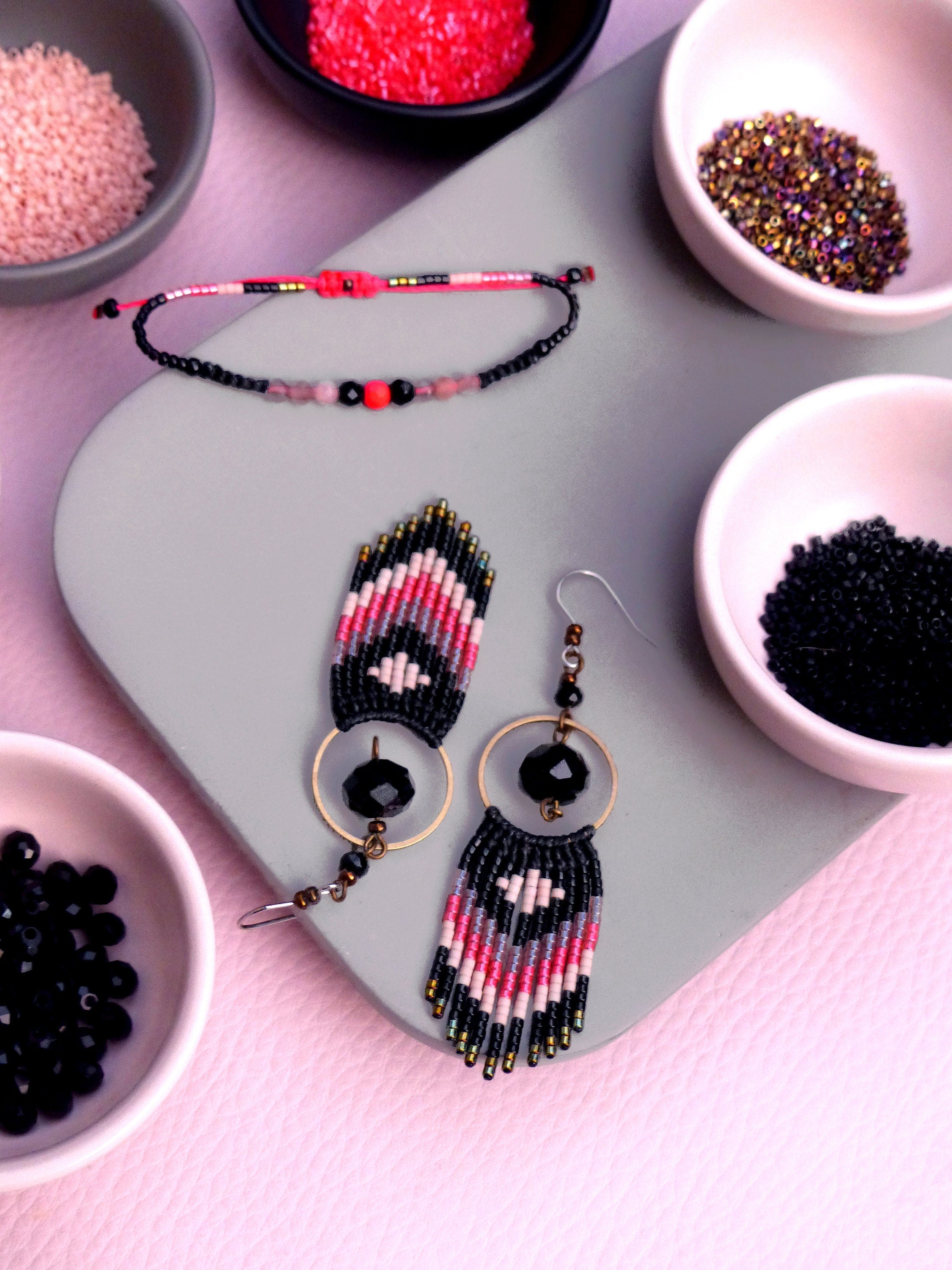Peut inclure: Une paire de boucles d'oreilles en perles noires, roses et blanches. Les boucles d'oreilles sont suspendues à des anneaux dorés avec des accents de cristal noir. Les boucles d'oreilles sont présentées sur une surface grise avec de petits bols de perles de différentes couleurs.
