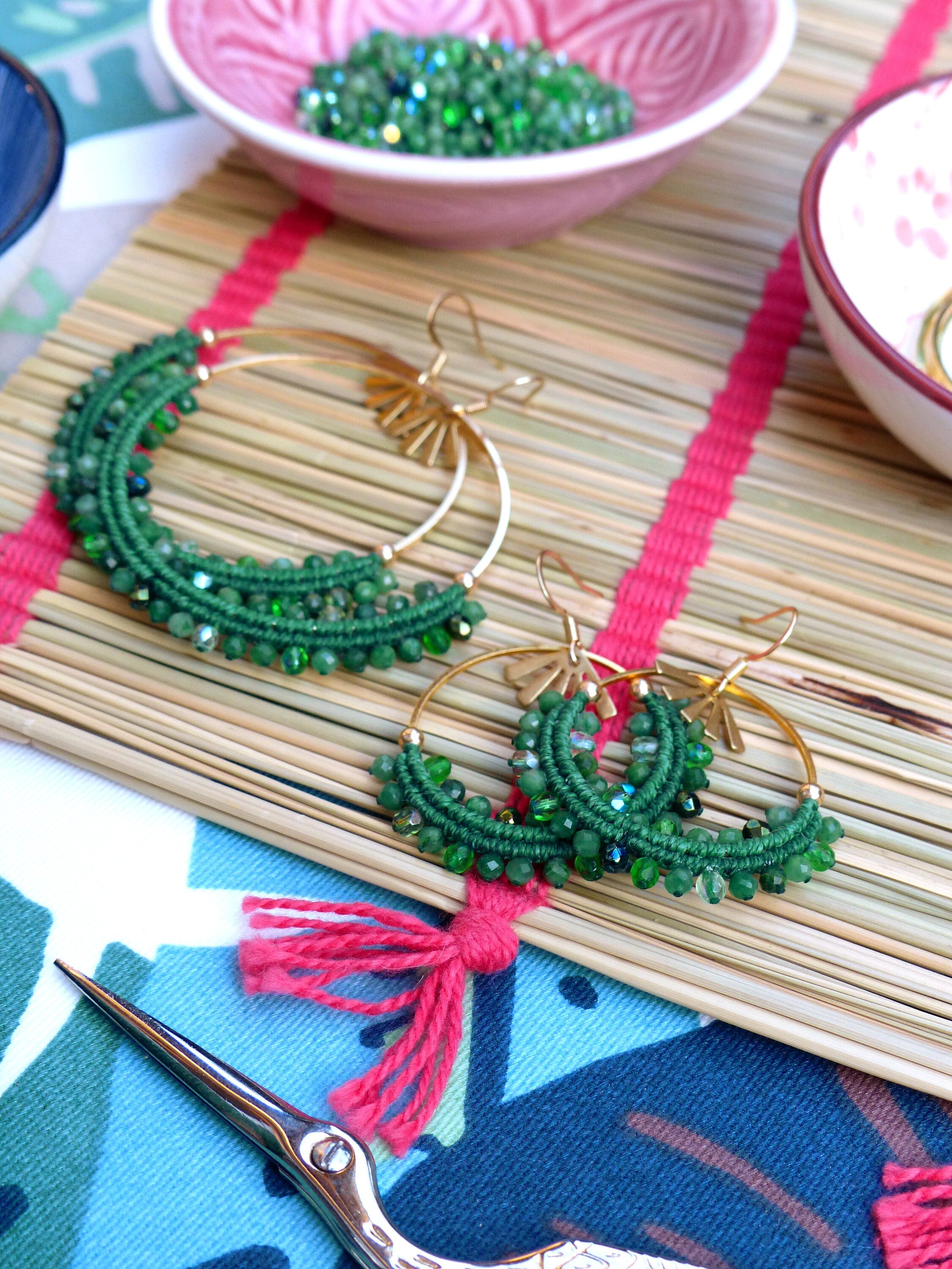 Peut inclure: Une paire de boucles d'oreilles créoles dorées avec des franges de perles vertes. Les boucles d'oreilles sont sur un tapis en bambou tissé avec un ruban rouge.