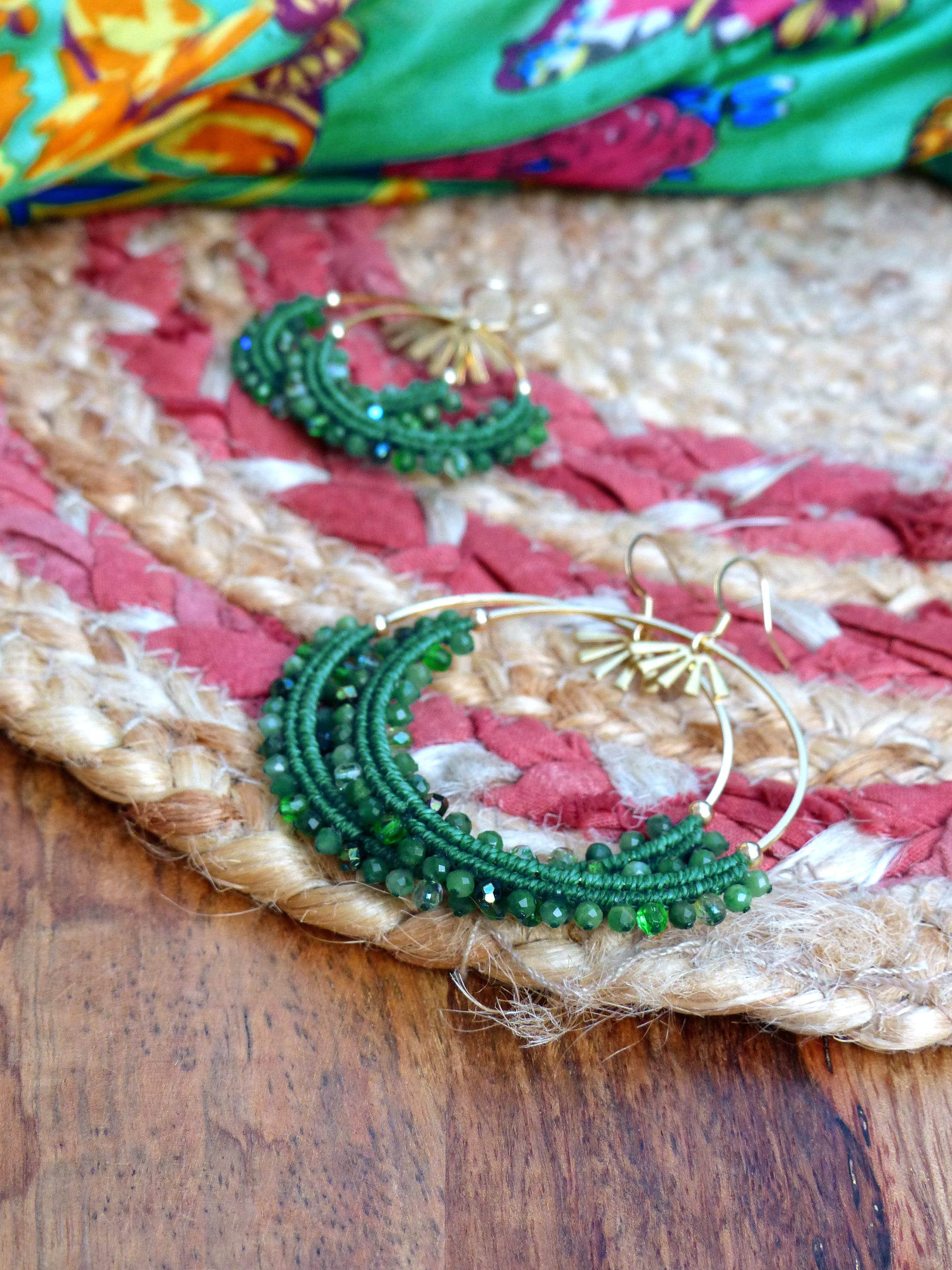 Peut inclure: Une paire de boucles d'oreilles créoles vertes et dorées avec un design perlé. Les boucles d'oreilles sont sur un tapis de paille tissé.