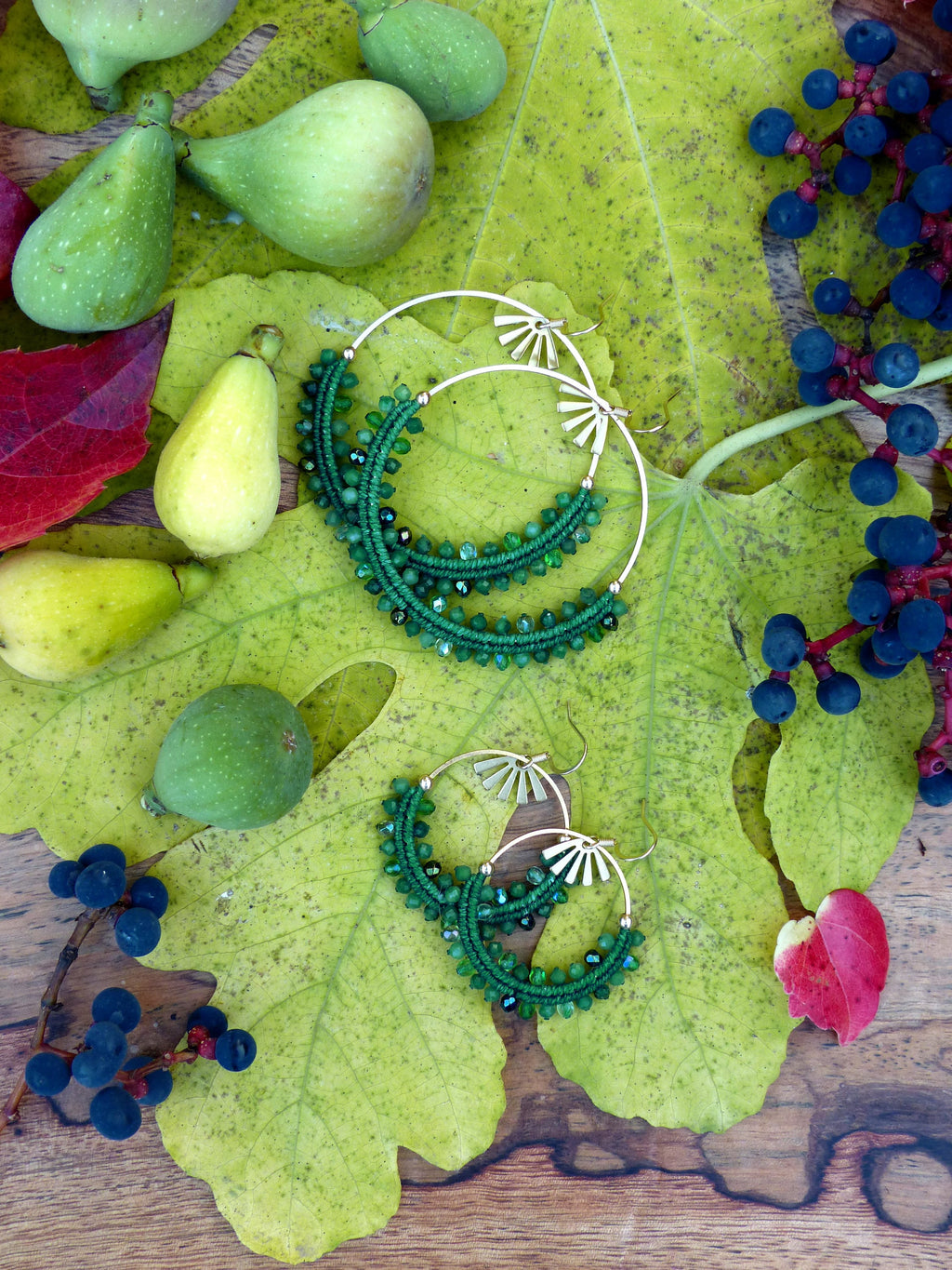 Peut inclure: Une paire de boucles d'oreilles créoles en perles vertes avec des accents dorés. Les boucles d'oreilles sont photographiées sur une feuille verte avec d'autres objets verts et bruns.