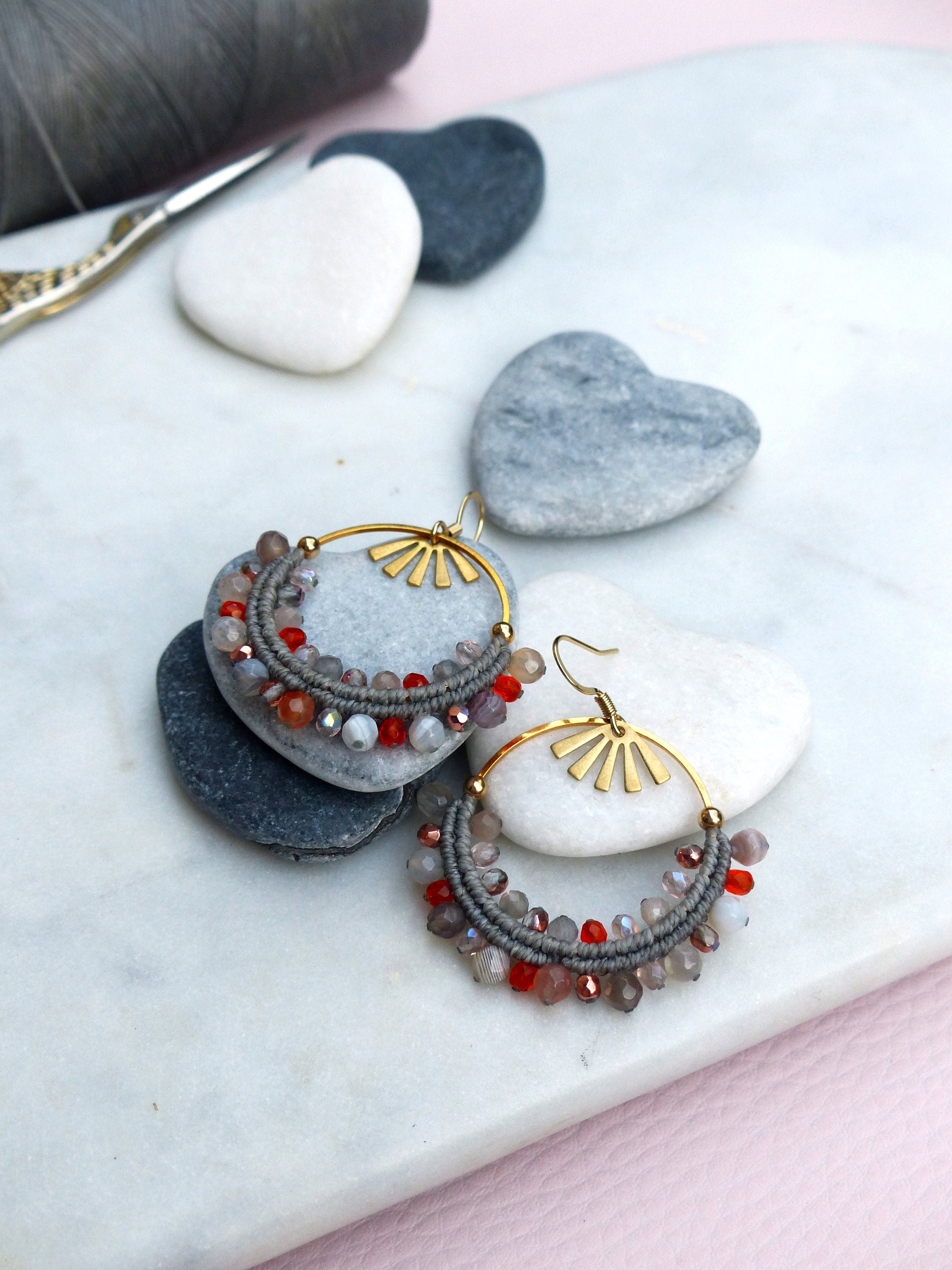 Peut inclure: Une paire de boucles d'oreilles créoles dorées avec des perles grises et rouges. Les boucles d'oreilles sont sur une surface blanche avec des pierres en forme de cœur grises.