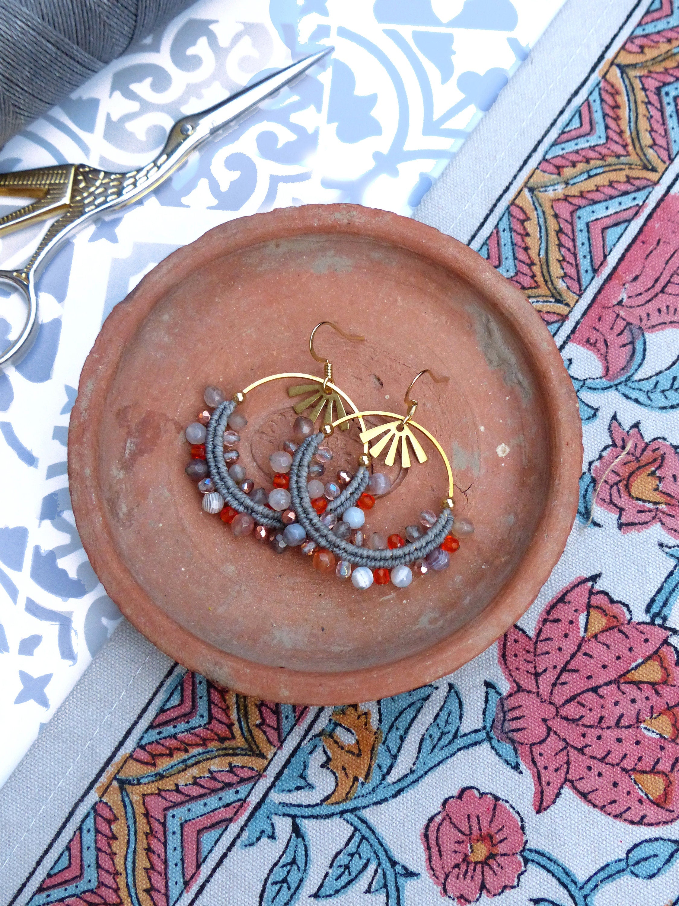 Peut inclure: Une paire de boucles d'oreilles créoles dorées avec des perles grises et orange. Les boucles d'oreilles sont sur une assiette en terre cuite.