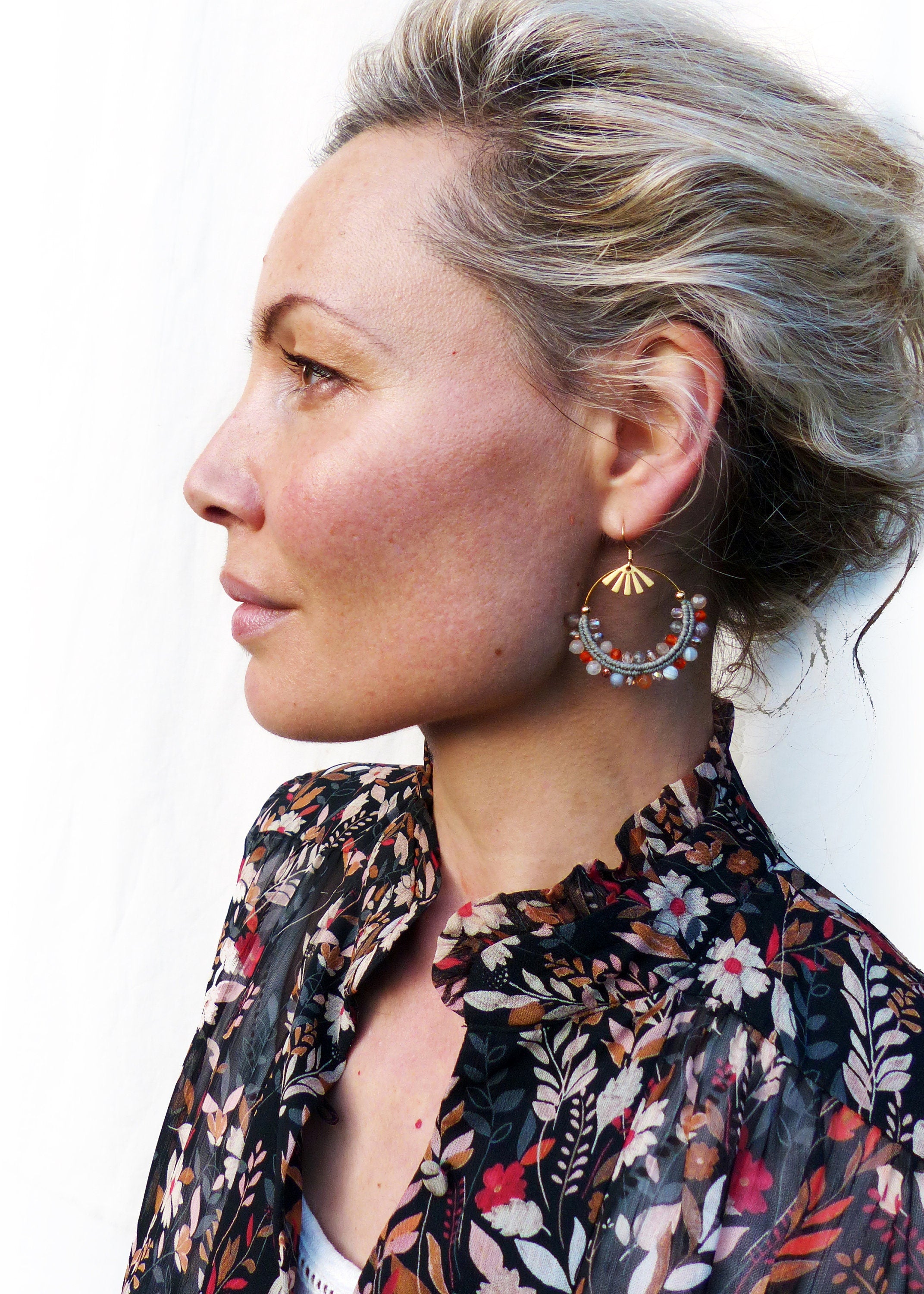 Peut inclure: Une femme portant un chemisier à imprimé floral avec des boucles d'oreilles créoles dorées ornées d'un motif en éventail et de perles colorées.