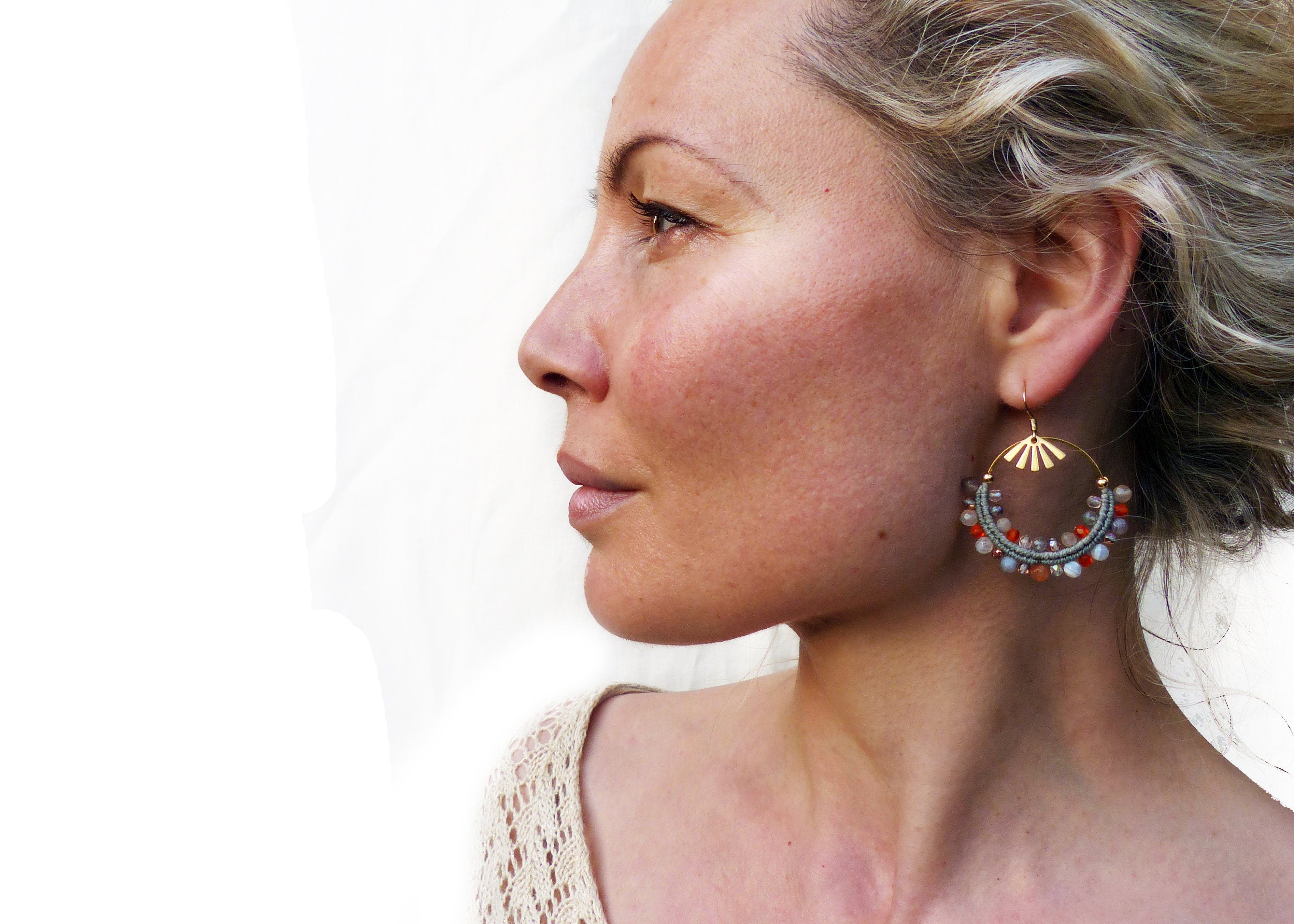 Peut inclure: Une paire de boucles d'oreilles créoles dorées avec un design perlé. Les perles sont dans des tons de gris, orange et rouge. Les boucles d'oreilles pendent de l'oreille d'une personne aux cheveux blonds.
